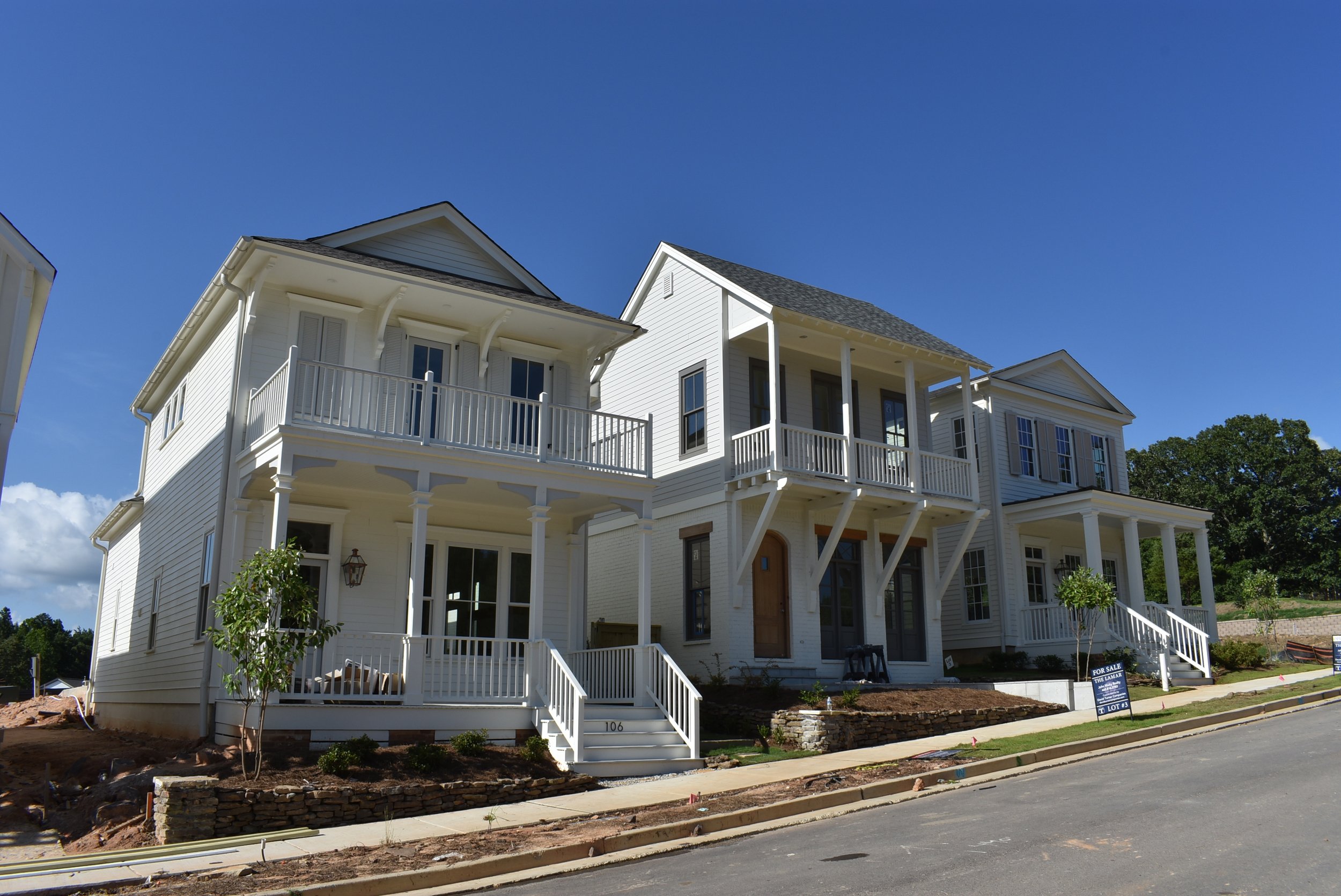 Newly constructed white multi-story house with balconies, front porch, and wooden stairs. The house has large windows and is set against a clear blue sky with some trees in the background.