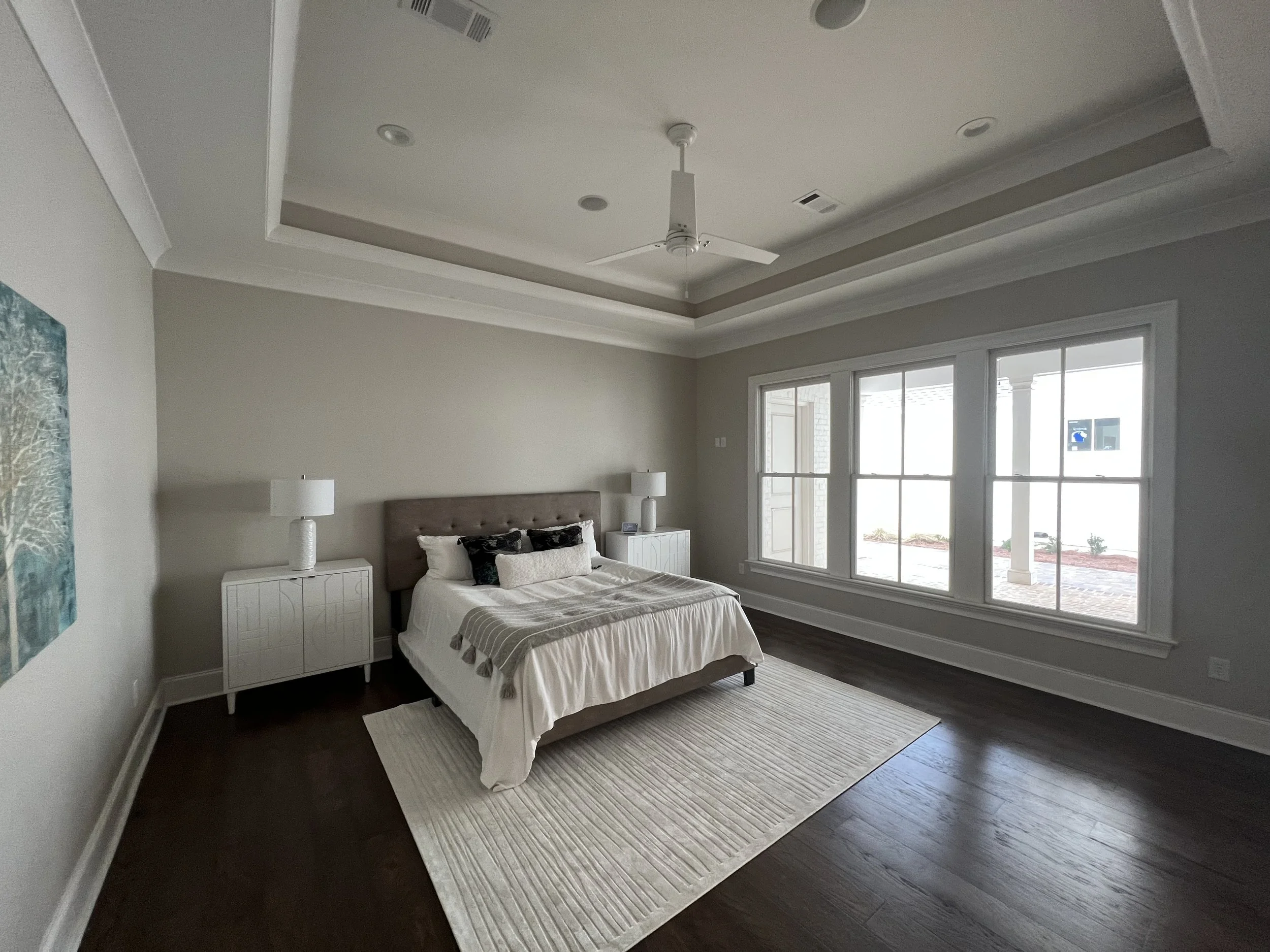 A bedroom with a bed, two white nightstands with lamps, a beige headboard, a large window, and hardwood floors.