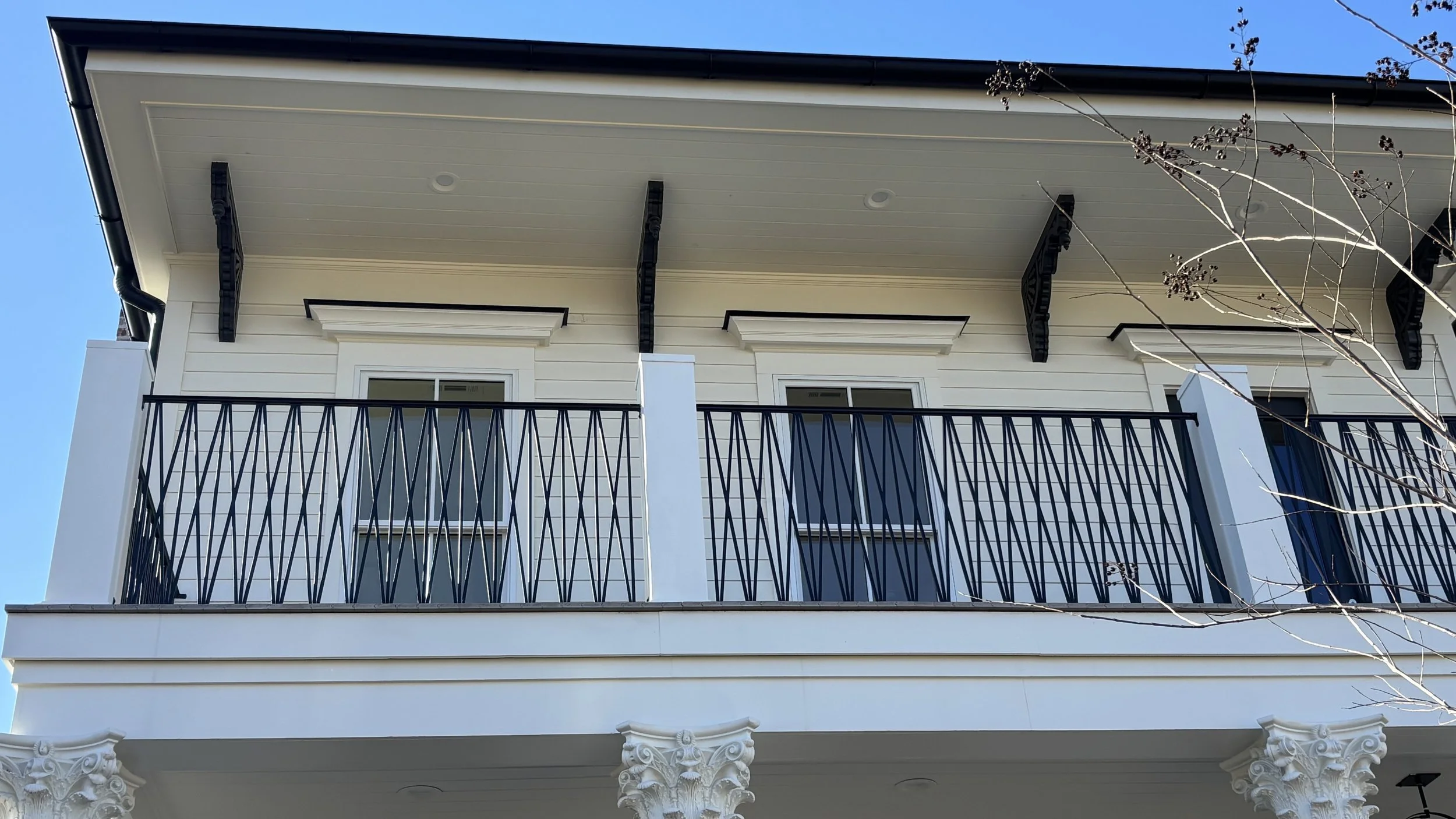 Close-up view of the upper part of a white building with decorative columns, two windows, and a black railing on a balcony. Tree branches are visible in the foreground. Clear blue sky in the background.