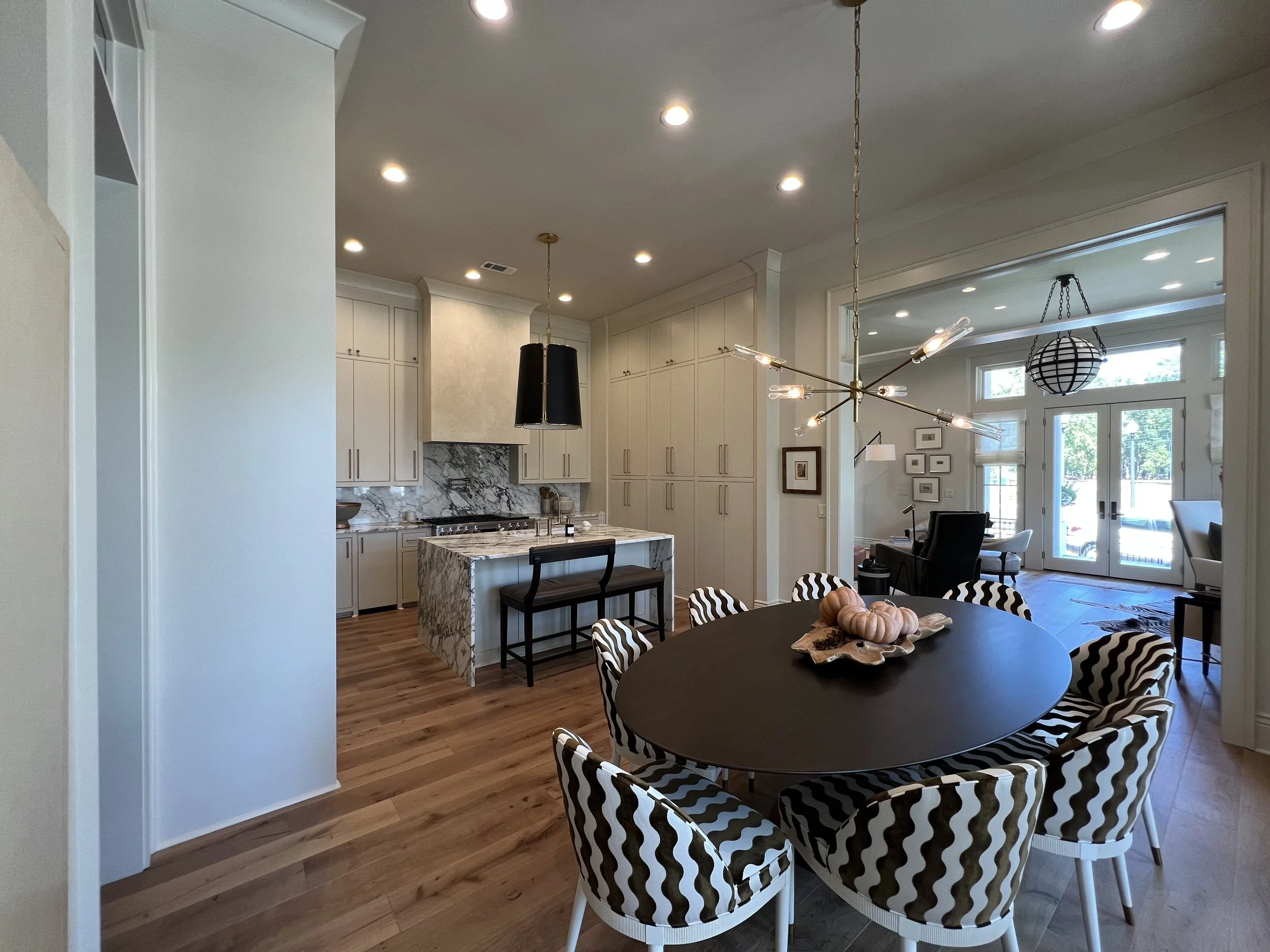 Open-concept kitchen and dining area with oak flooring, white cabinetry, marble kitchen island, black pendant lights, modern chandelier, and a dining table with black and white zigzag-patterned chairs.
