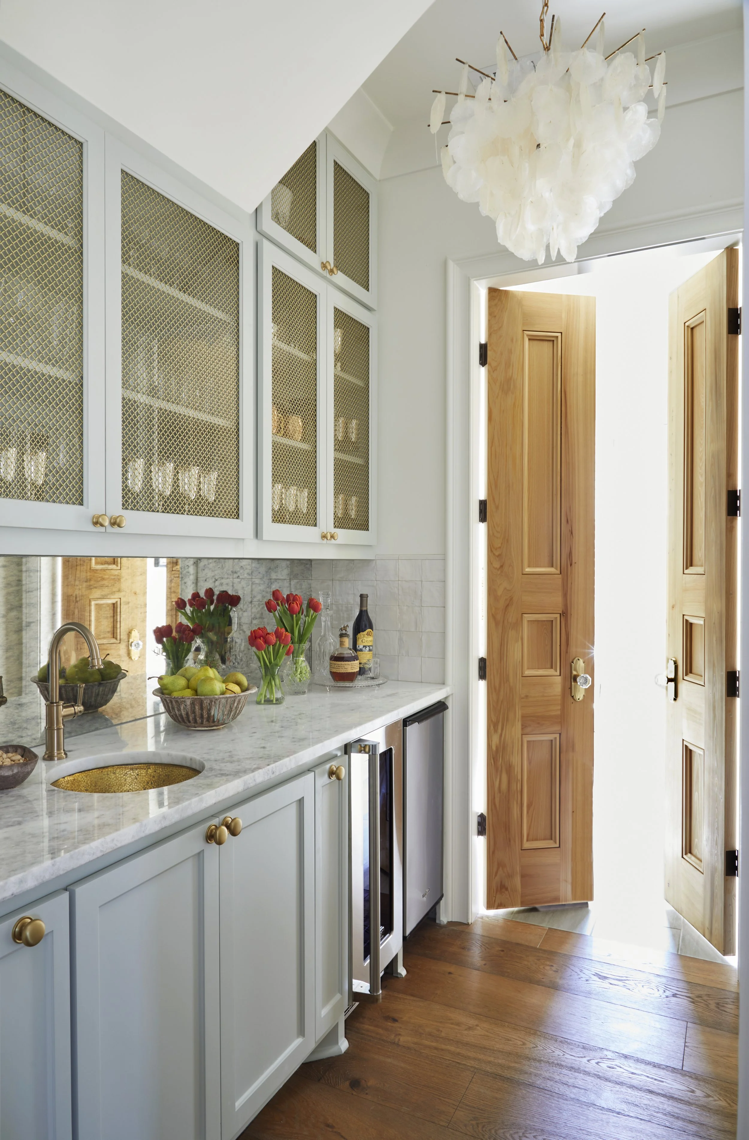 Bright kitchen with light blue cabinets, marble countertop, wooden doors, a floral chandelier, and fresh flowers on the counter.