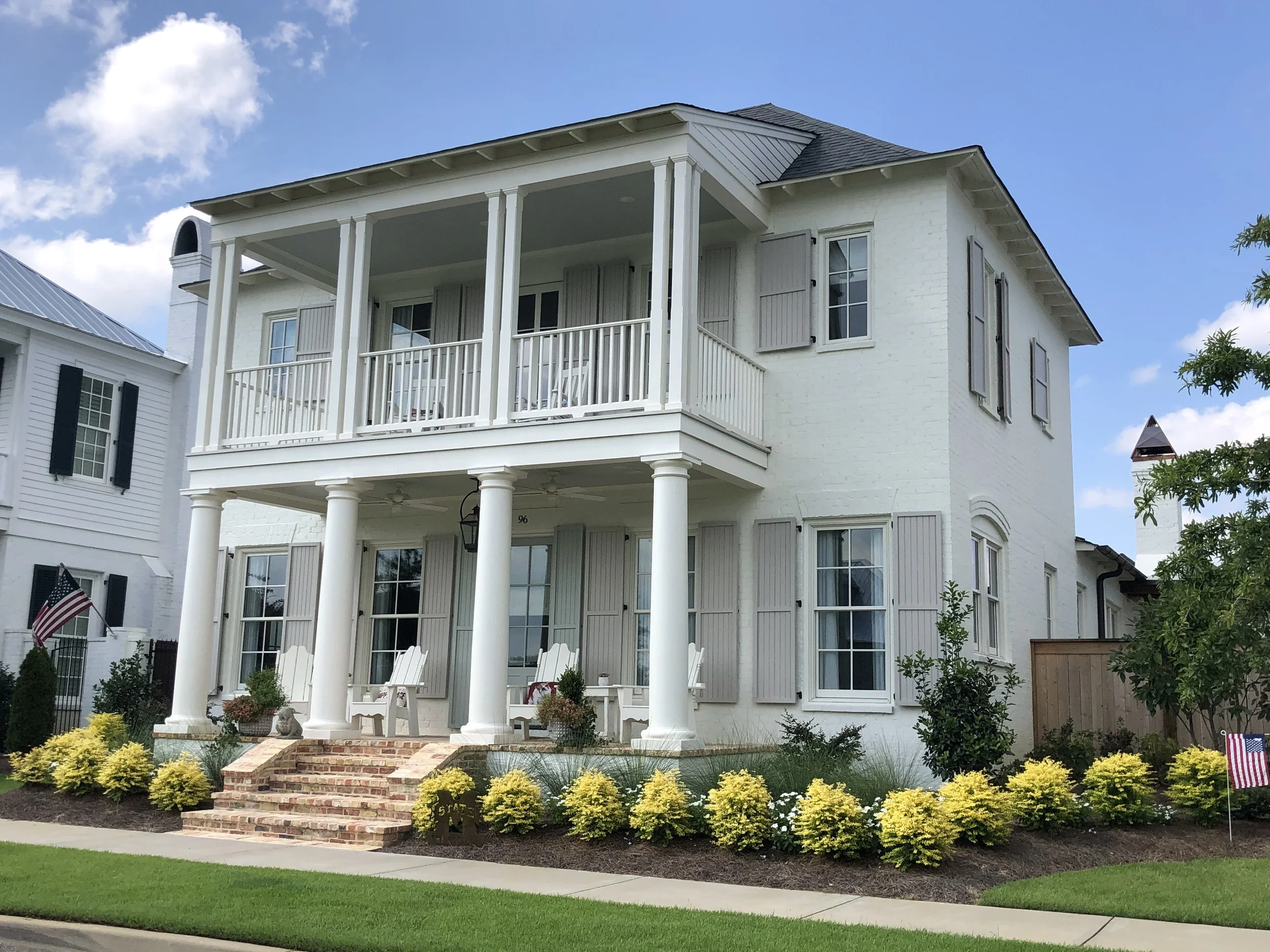White two-story house with columns, front porch, and balcony, surrounded by landscaped yard with yellow bushes and American flags, under a blue sky with clouds.