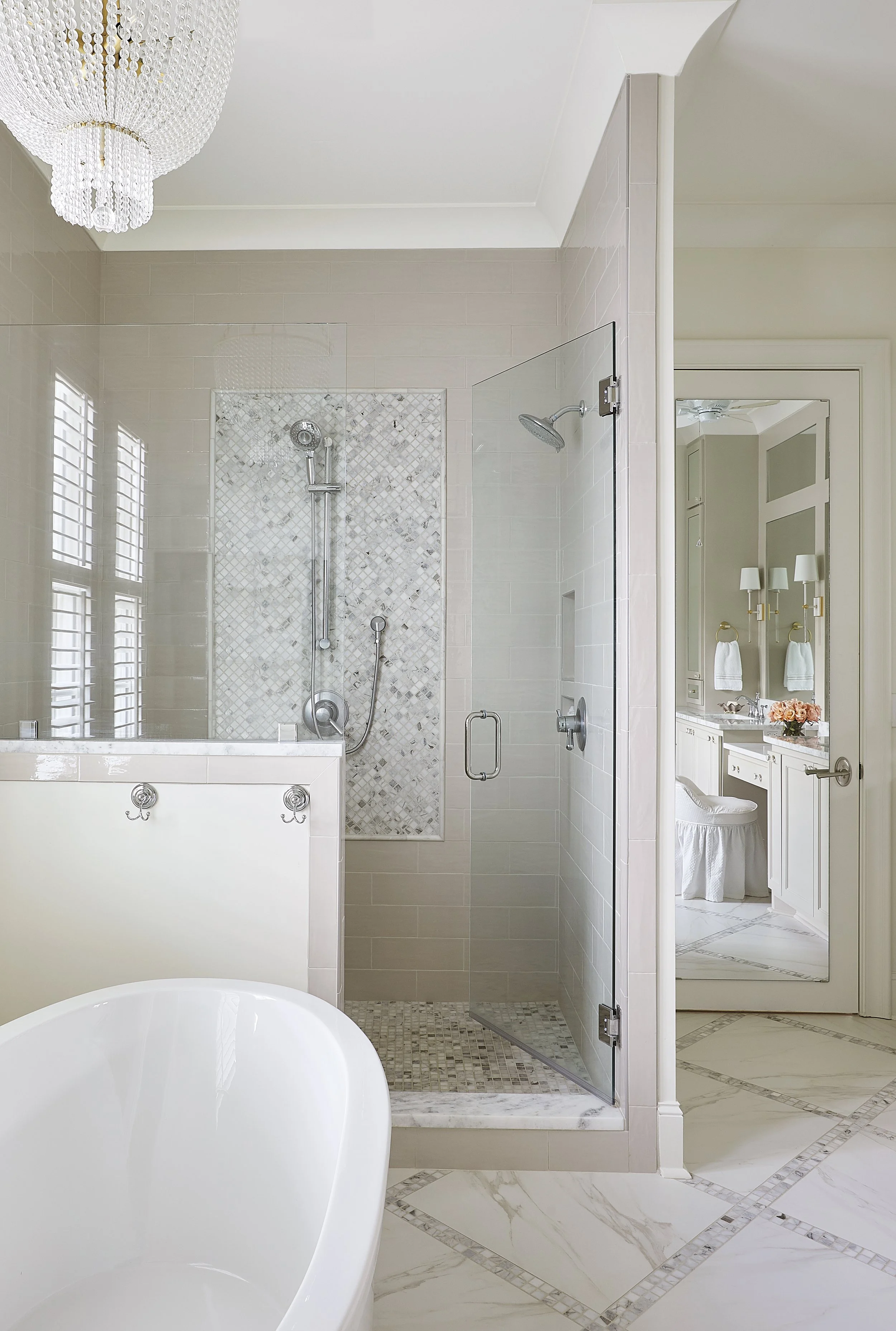 A luxurious bathroom with a white freestanding bathtub, a glass-enclosed shower with grey and white mosaic tile accents, beige tiled walls, and a white vanity with a mirror and wall-mounted lights.