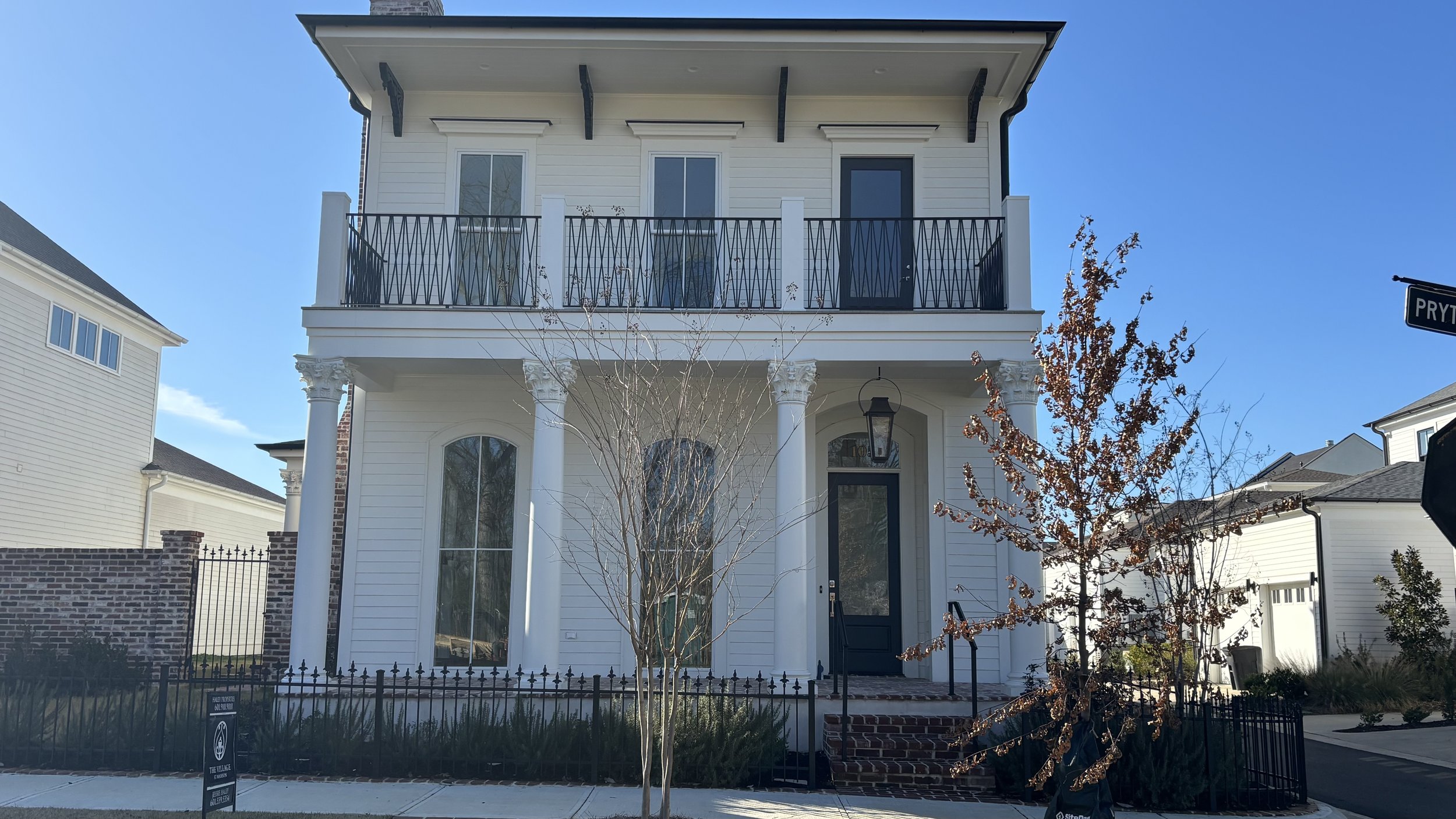 White two-story house with black door, columns, a balcony with black railing, and a small front yard with a tree, a black fence, and a brick walkway.