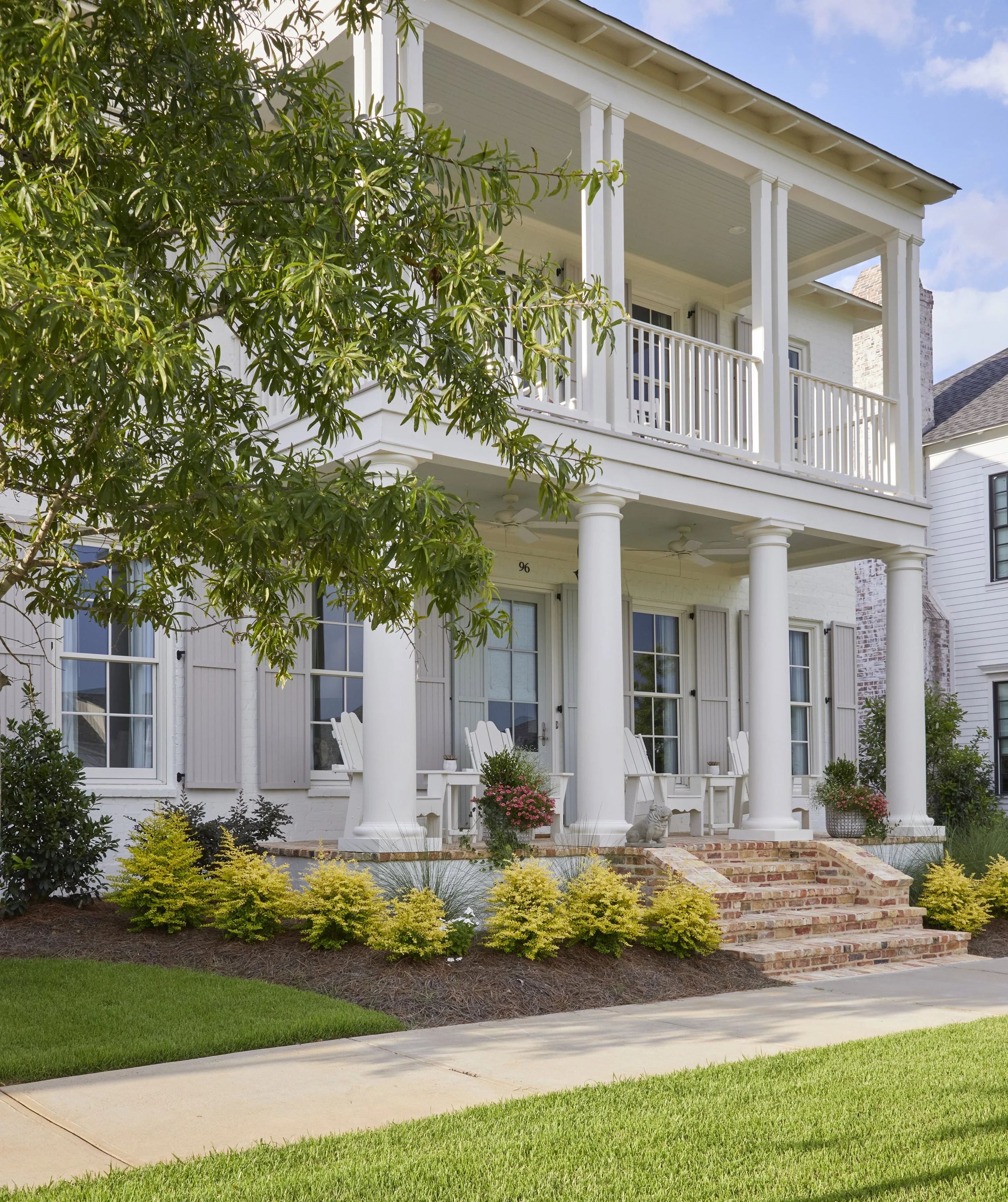 Two-story white house with a porch and columns, brick stairs, green lawn, trees, and outdoor chairs.