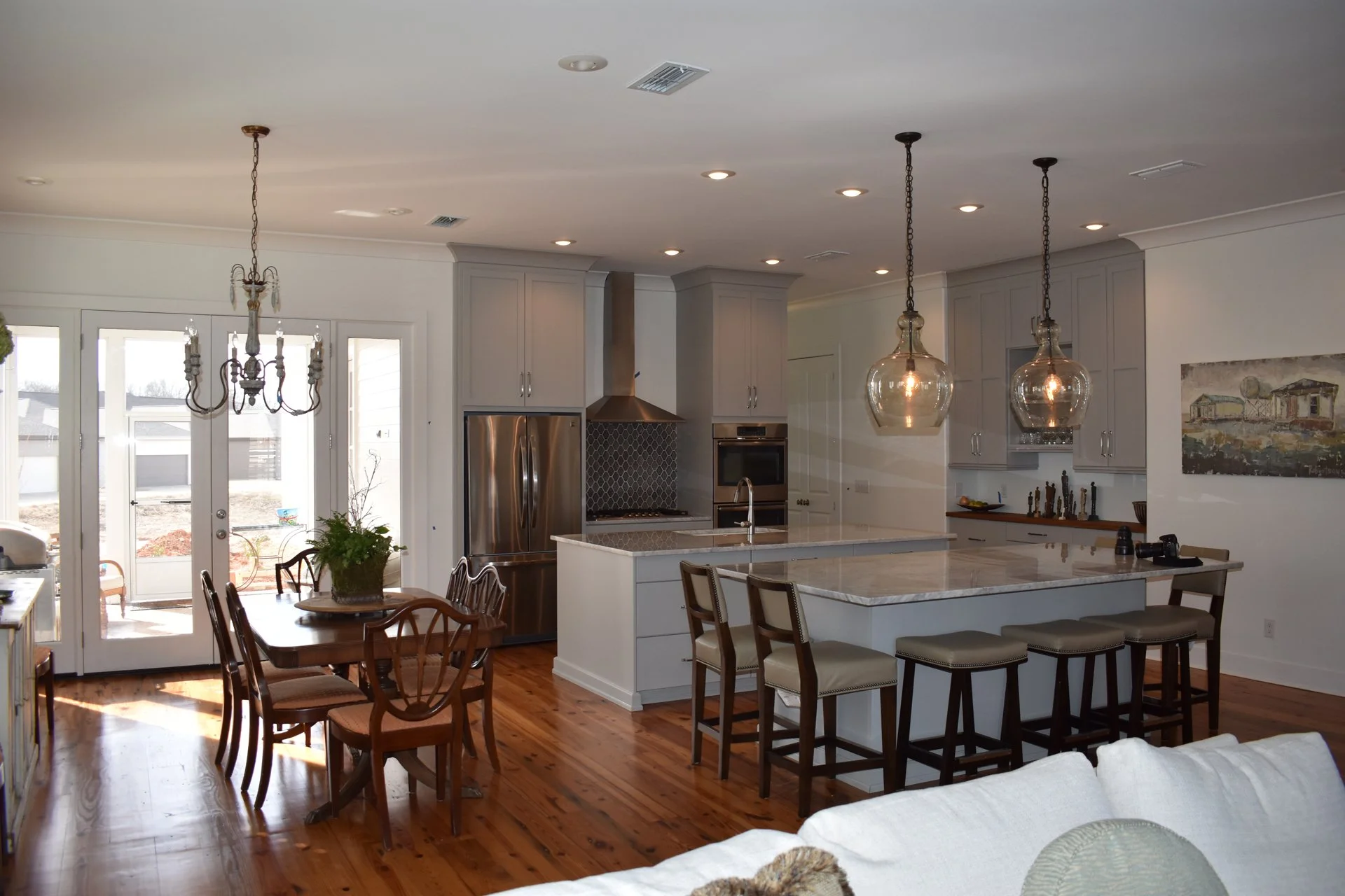 Open-concept kitchen and dining area with white cabinets, stainless steel appliances, a marble island with bar stools, wooden dining table, pendant and chandelier lighting, hardwood floors, and large glass doors leading outside.