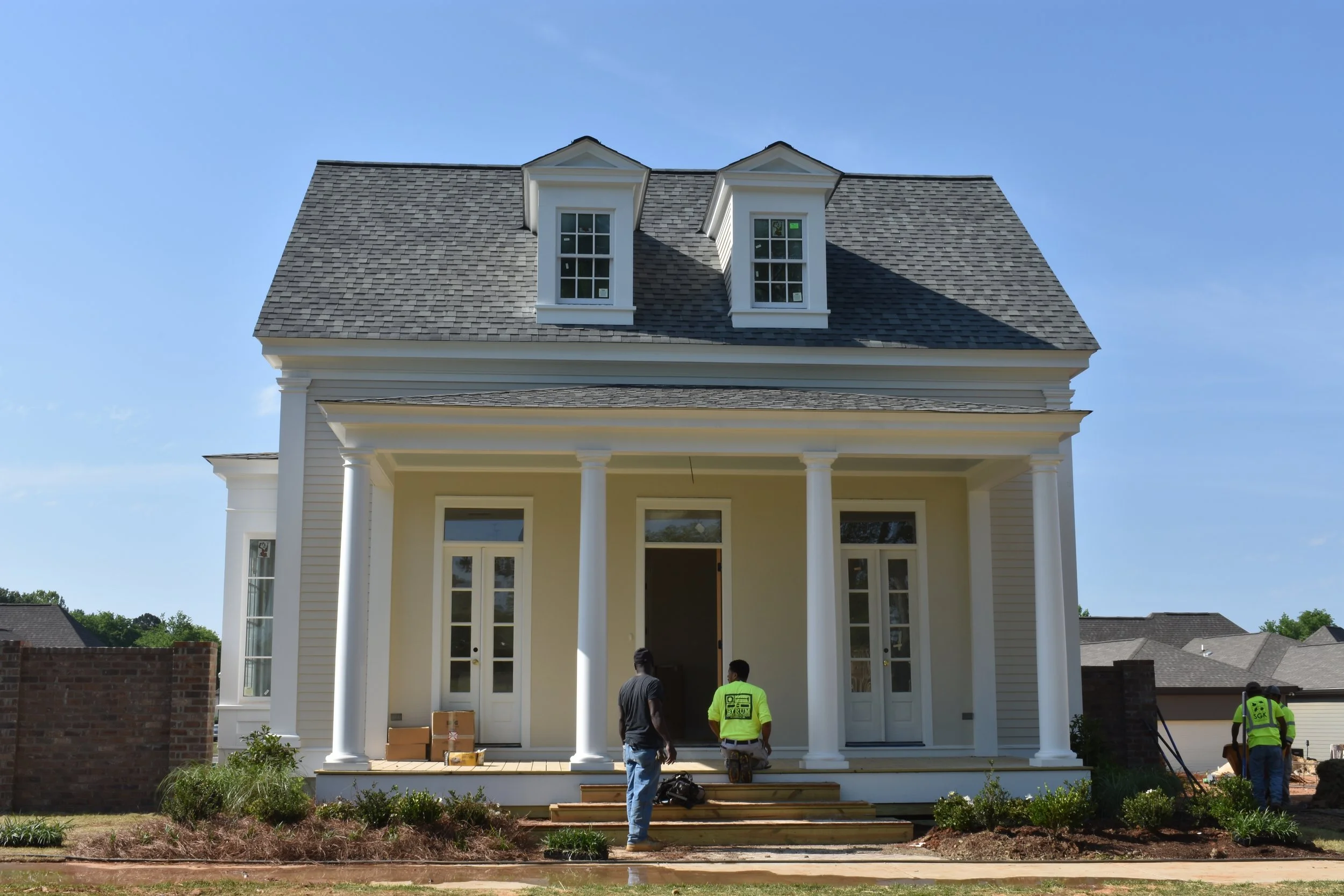 Construction workers building a large, white, two-story house with a porch and columns. The house has two dormer windows on the roof and a front door with glass panels. The scene includes grass, shrubs, and a clear blue sky.