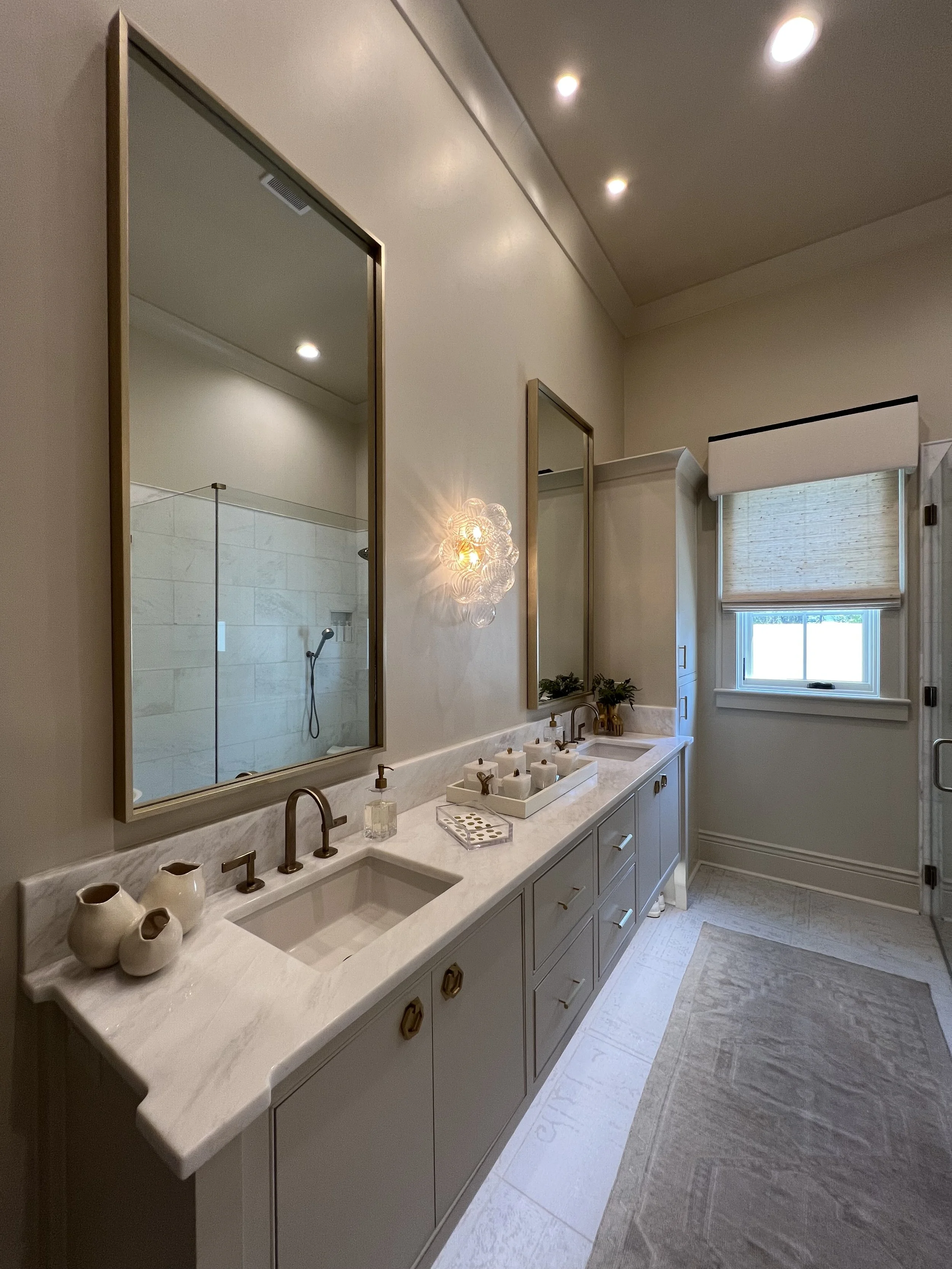 Modern bathroom with dual sinks, large mirrors, beige cabinetry, decorative vases, and a window with blinds.