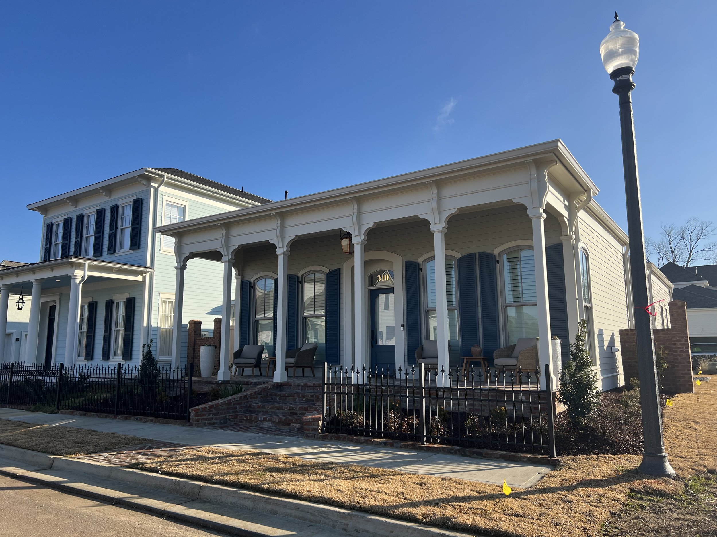 A two-story house with a front porch, white siding, blue shutters, and outdoor seating, next to a street with a lamp post.
