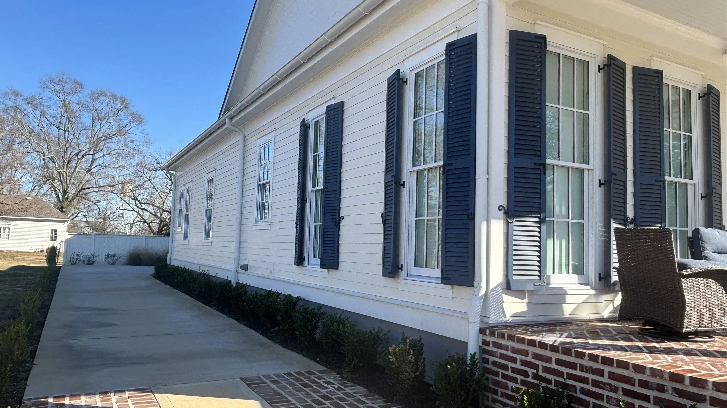 Exterior of a white house with black shutters and a brick patio with outdoor furniture under a blue sky.