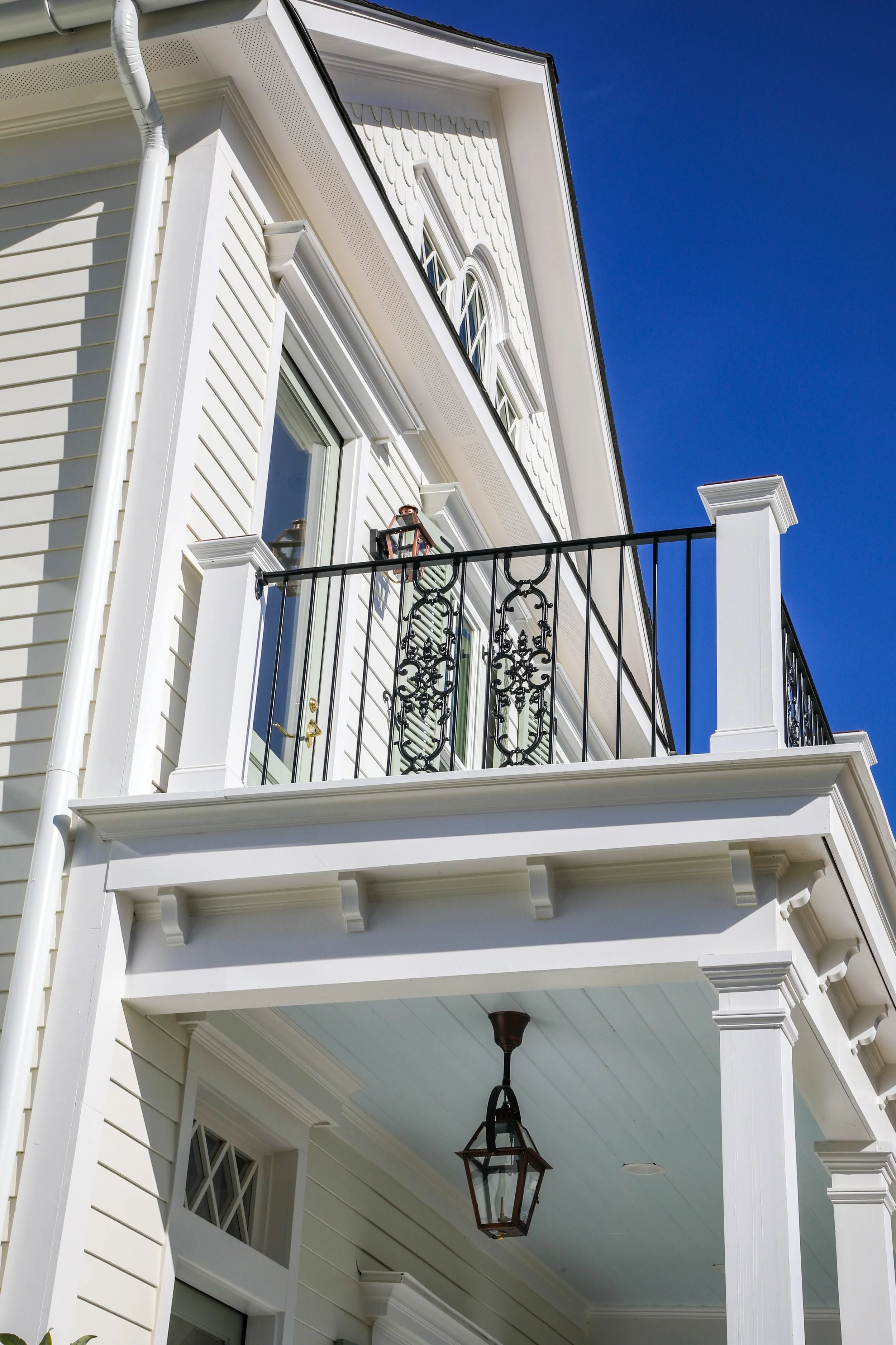 Close-up of a white house with a balcony, black wrought iron railing, a lantern on the balcony, and a hanging lantern in the porch, against a clear blue sky.