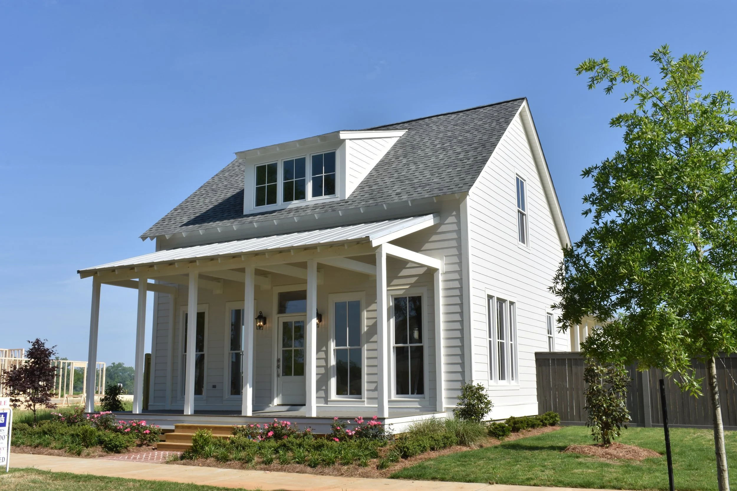 White two-story house with a porch, gray roof, and multiple windows, surrounded by landscaped garden and trees.