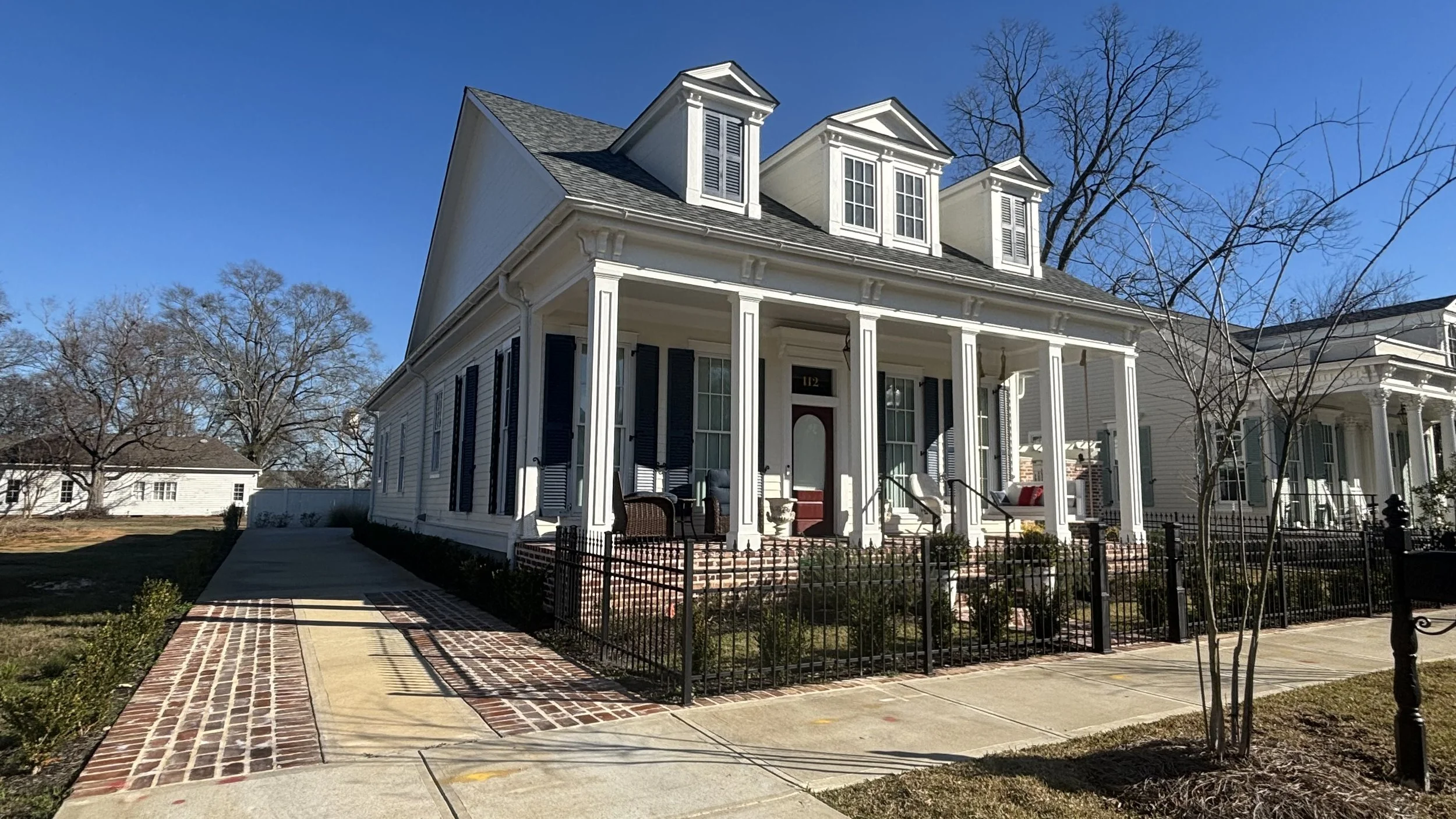 White two-story house with a porch, black shutters, and dormer windows, surrounded by a black fence and a brick and concrete sidewalk in the foreground.