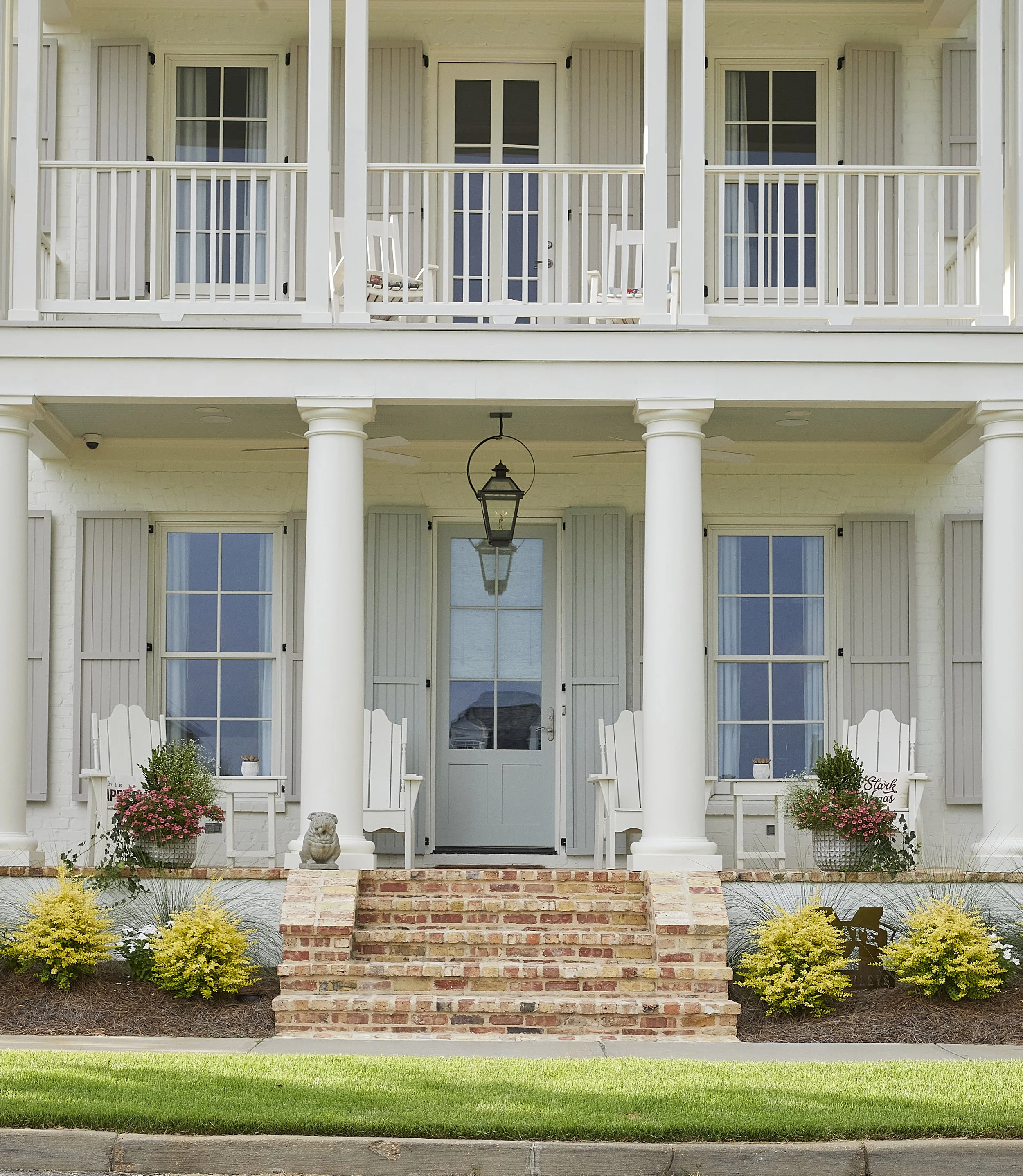 Front porch of a white two-story house with brick stairs, potted flowers, white chairs, and a lantern hanging from the ceiling.