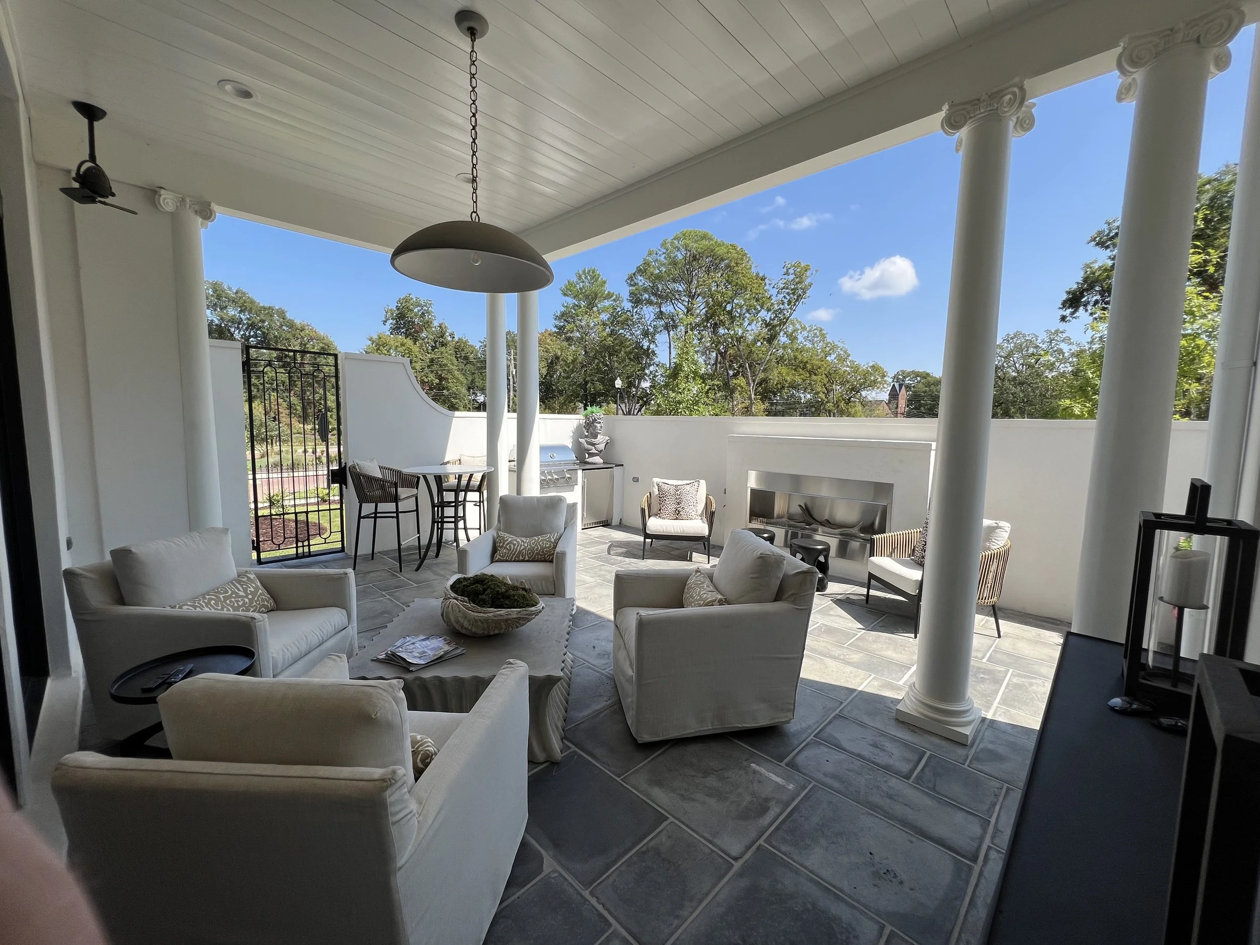 Covered outdoor patio with white cushioned seating, a stone fireplace, a bar table with chairs, and a barbecue grill, overlooking trees and a bright blue sky.