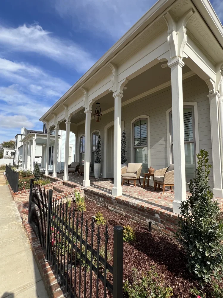 Front porch of a house with white columns, chairs, potted plants, brick walkway, a small black fence, and a landscaped garden bed under a partly cloudy sky.