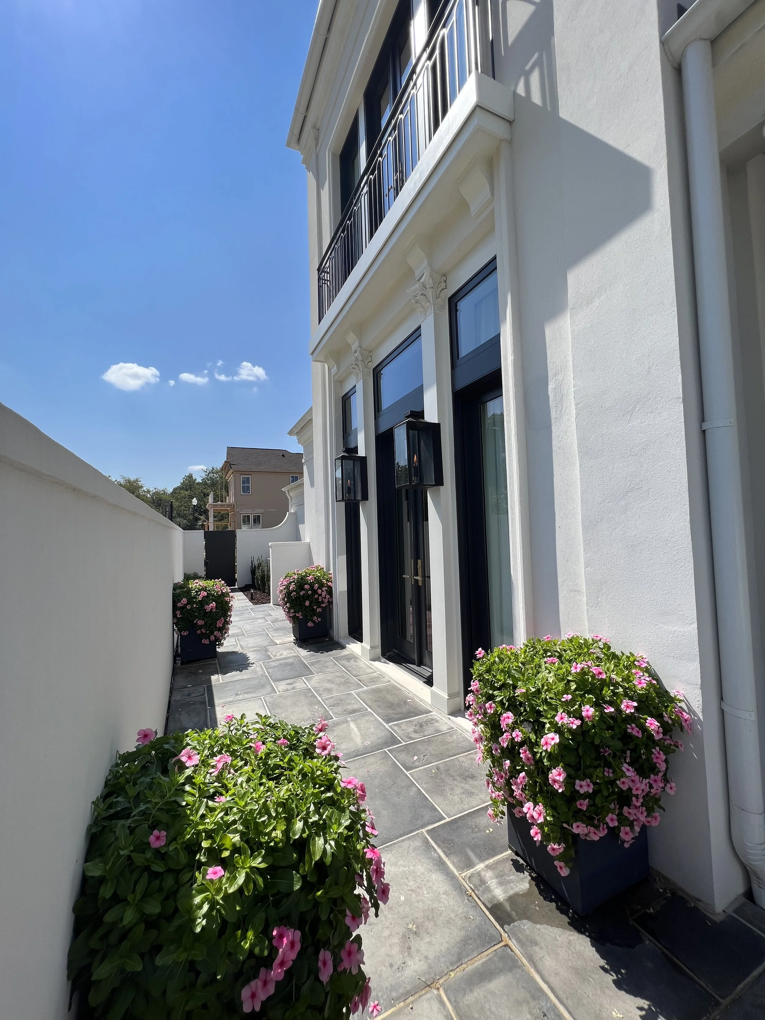 A narrow outdoor patio with potted pink flowers on either side, leading to a black door with glass panels, attached to a white brick building under a bright blue sky.