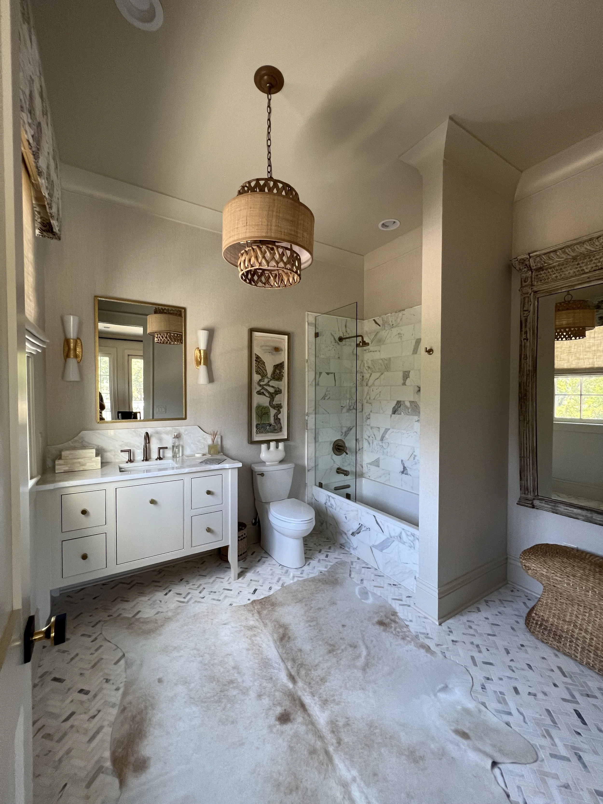 Bathroom with white vanity, mirror, framed wall art, toilet, and shower with glass door and marble tiles, wicker bench, and rug.