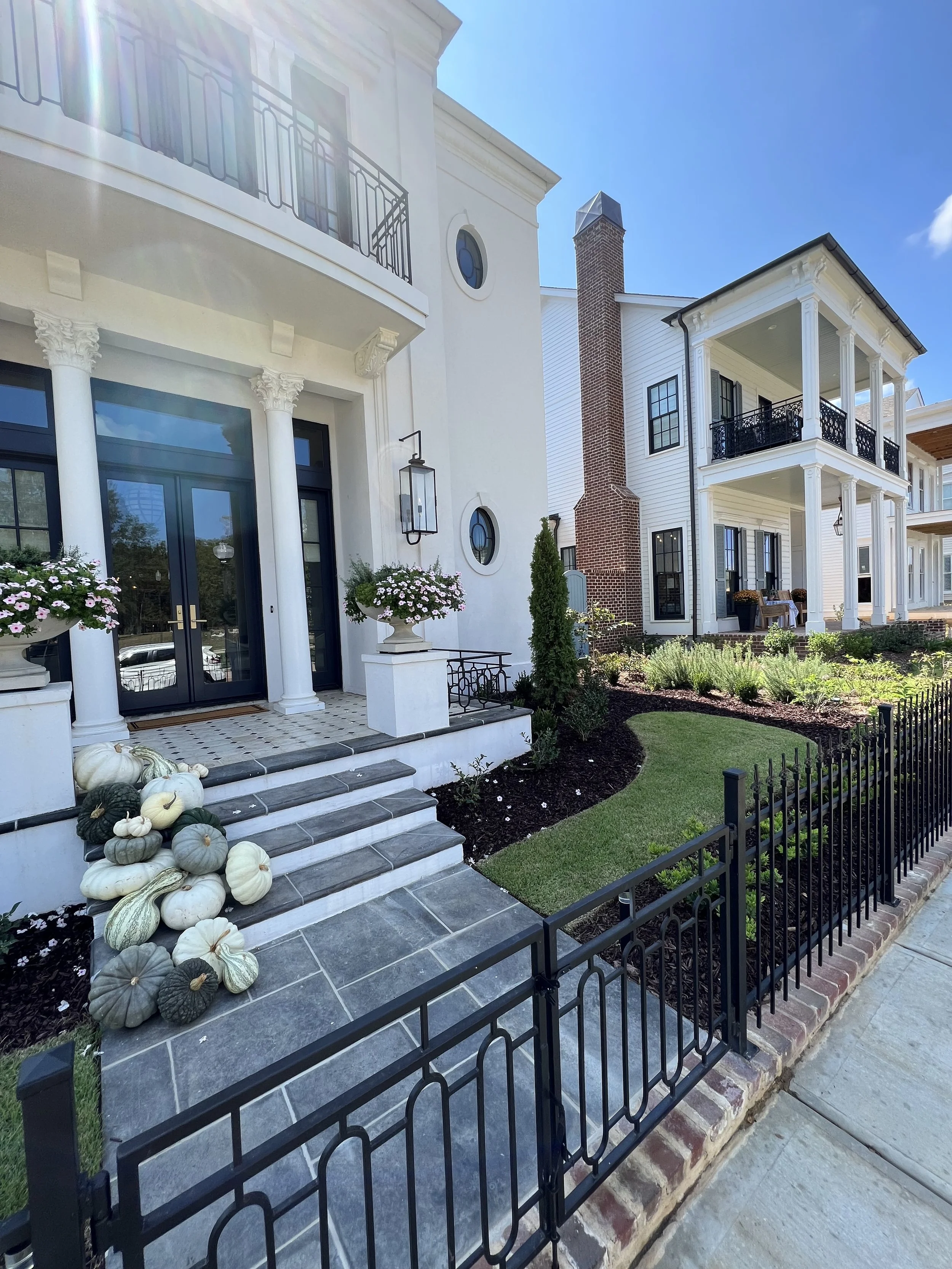 Front view of a white house with black and grey details, featuring columns, round windows, and a brick chimney. The house is decorated with pumpkins and gourds on the steps, and there are potted flowers and a small landscaped garden with green grass 