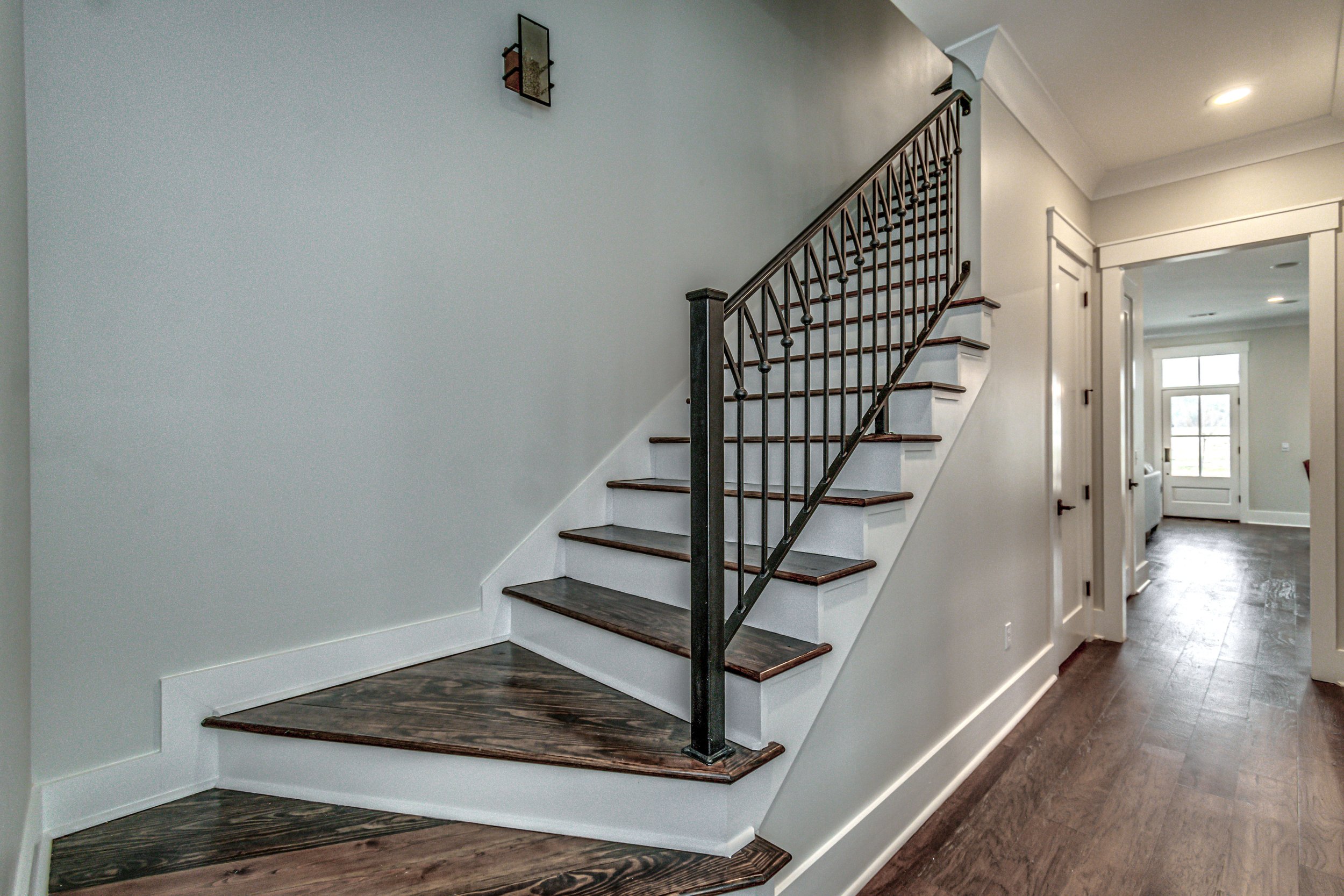Interior view of a home's staircase with dark wooden steps, black metal railing, and a light gray wall. There is a small decorative piece on the wall and a hallway leading to a bright room with a window.