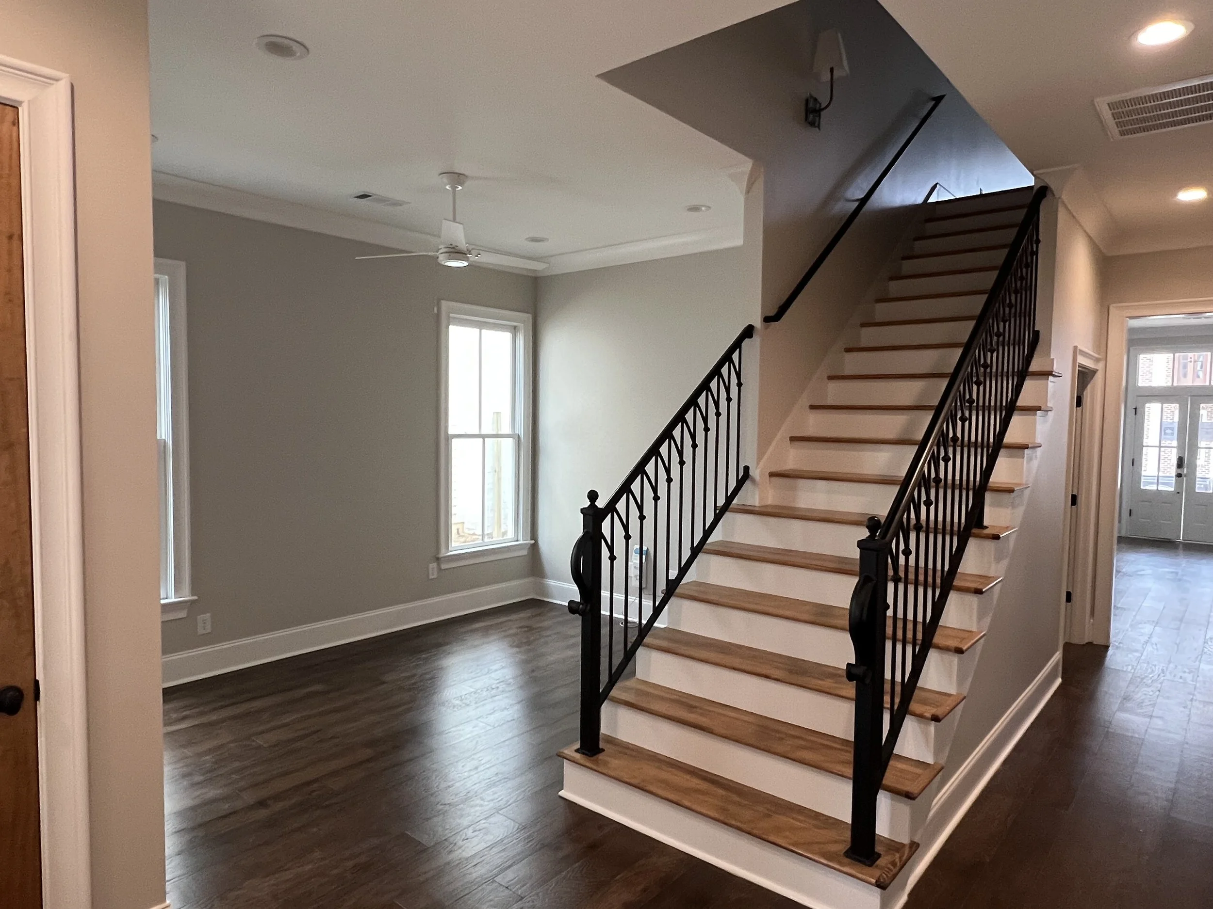 Empty living room with dark hardwood floors, white trim, large windows, a white ceiling fan, and a staircase with wooden steps and black metal railing.