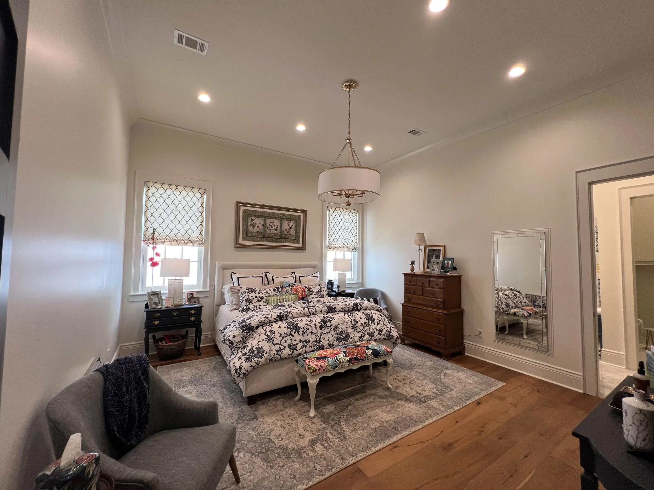 A bedroom with a bed, side tables with lamps, a dresser, a mirror, and decor. The room features hardwood flooring, a patterned rug, and two windows with blinds, creating a cozy and elegant atmosphere.