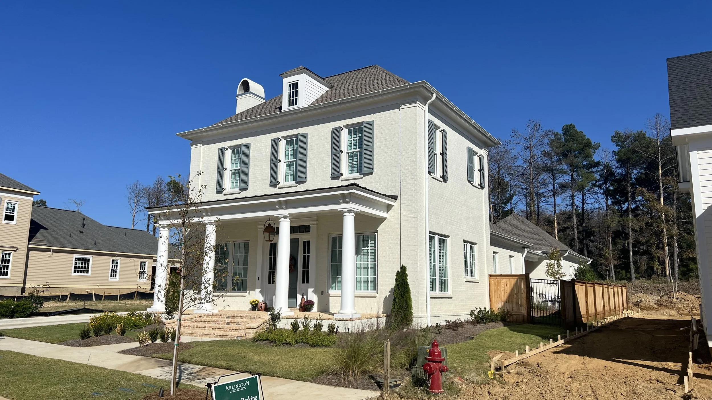 A white two-story house with black shutters, a front porch with columns, a landscaped yard, and neighboring homes, under a clear blue sky.