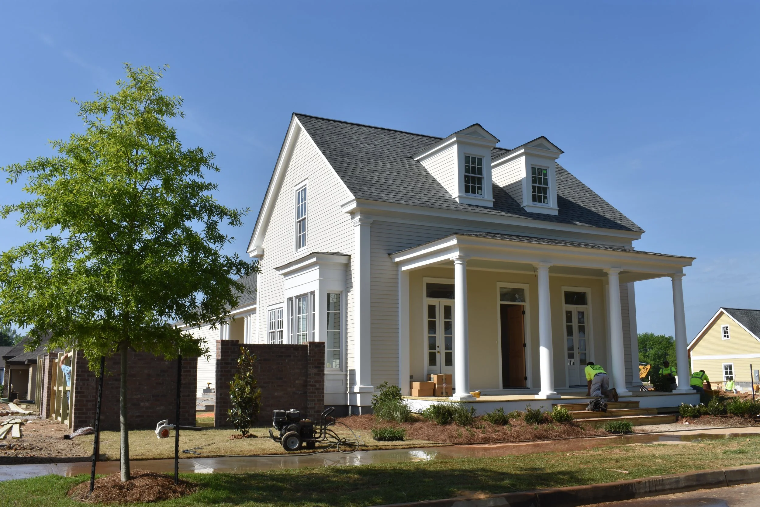 A white two-story house under construction with a front porch supported by columns, large windows, and a gray shingle roof. Workers in safety vests are working on the porch steps. There are trees, a brick wall, and construction equipment in the yard.