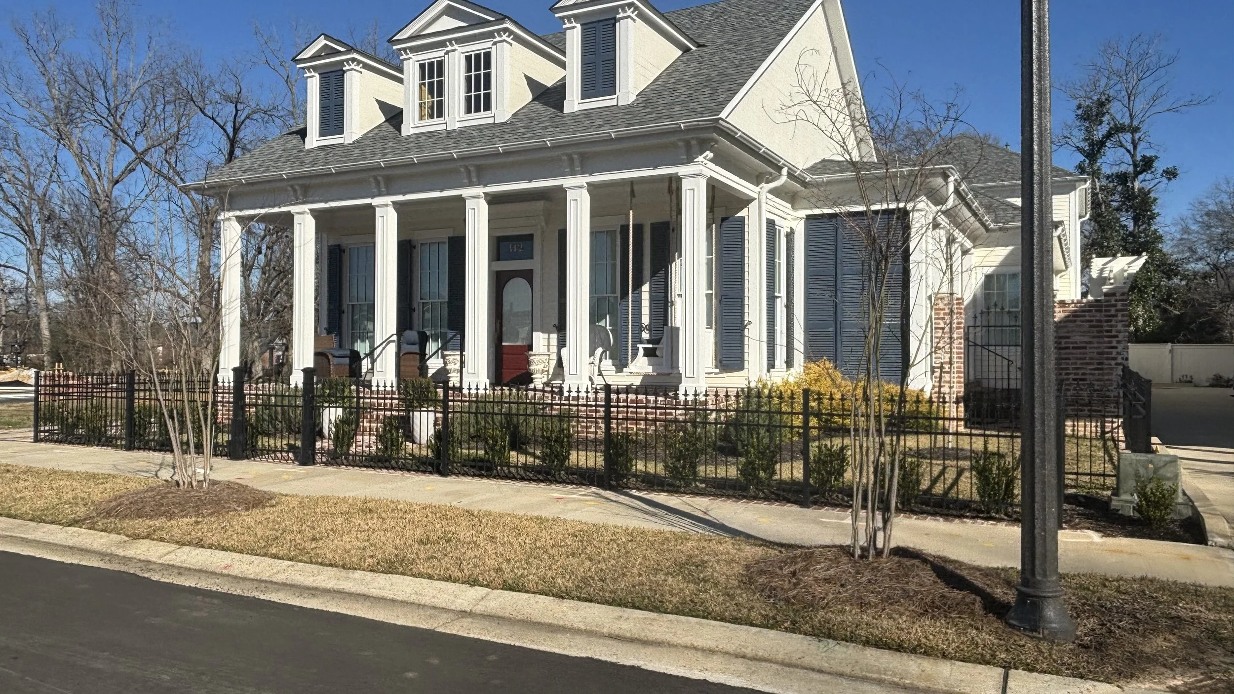 A large, elegant house with white columns, bay windows, and a front porch, surrounded by a black fence and a landscaped yard in a suburban neighborhood.