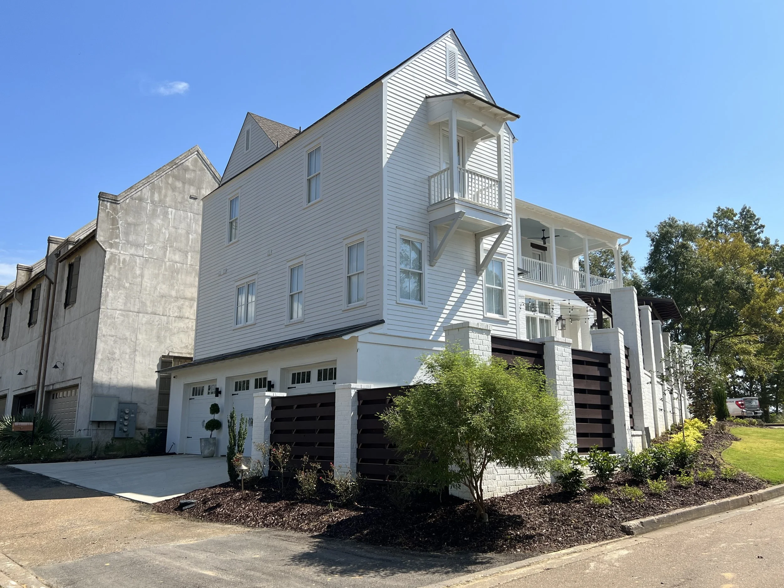 A modern, multi-story white house with balconies and a small front yard, located in a neighborhood with other houses and trees, under a clear blue sky.