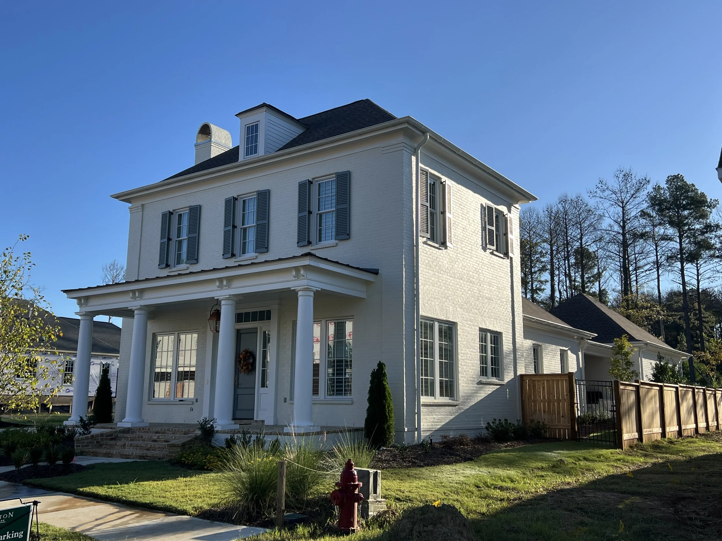 A white, three-story house with blue shutters, a porch with four white columns, a brick staircase, and a front yard with plants and a fire hydrant, under a clear blue sky.