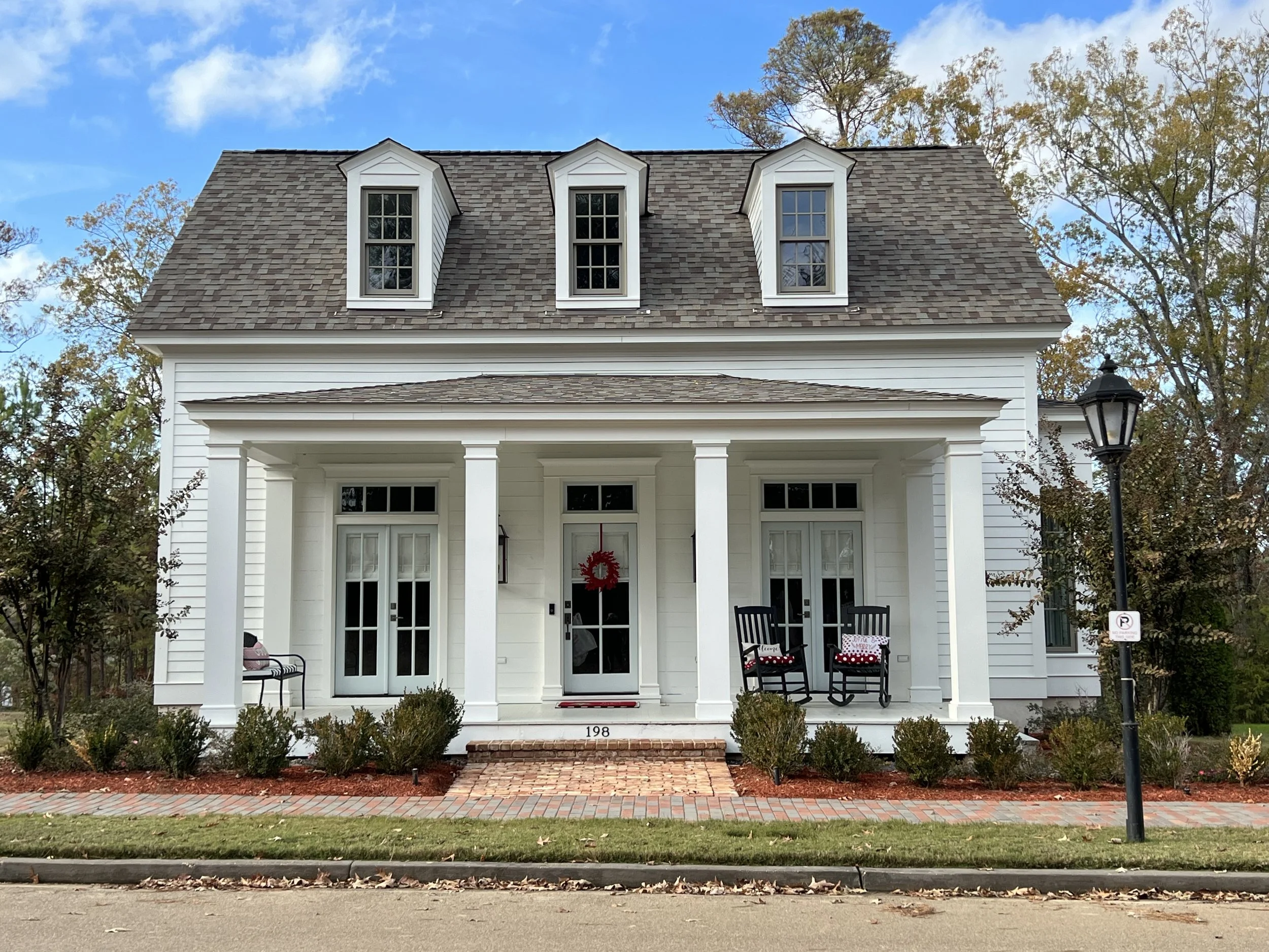 A white two-story house with a brick front porch, four black rocking chairs, and a red wreath on the door, surrounded by trees and a landscape with bushes and mulch, under a partly cloudy sky.