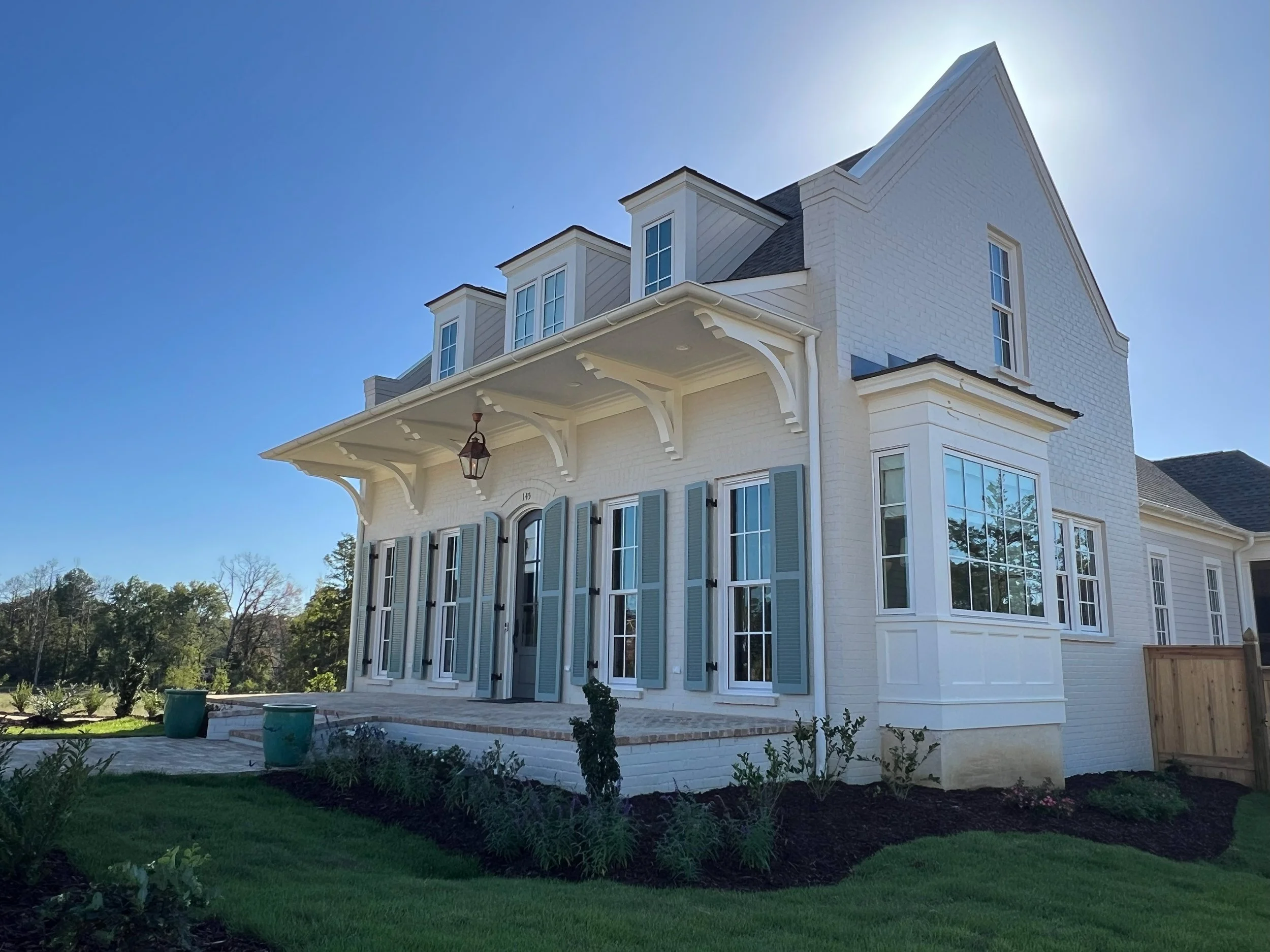 A large white house with multiple stories and blue shutters, featuring bay windows and a covered porch with decorative brackets, is set against a clear blue sky. The front yard includes a lawn, flower beds, and young plants.
