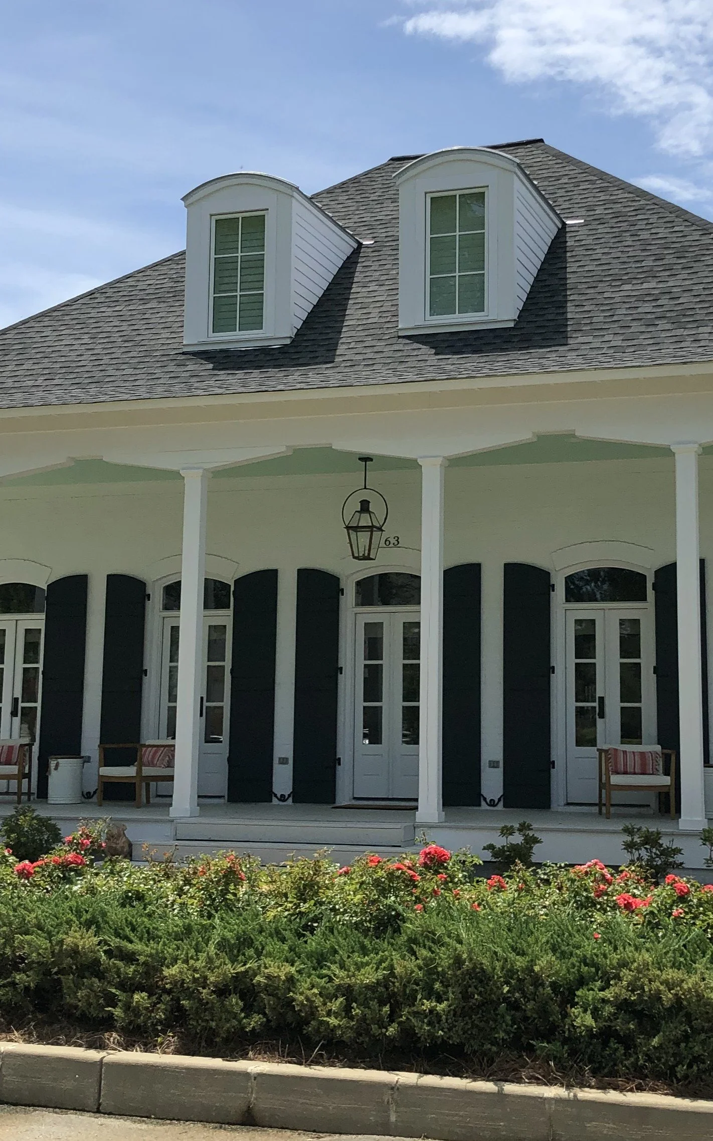 Front view of a house with a covered porch, black shutters, white doors, and a gray roof with two dormer windows. There are flower beds with pink flowers in the front and a sidewalk along the street.