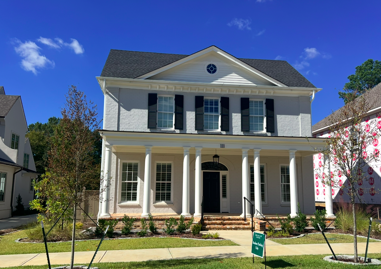 White two-story house with black shutters and six front columns, front porch, and brick steps in a neighborhood with trees and clear blue sky.
