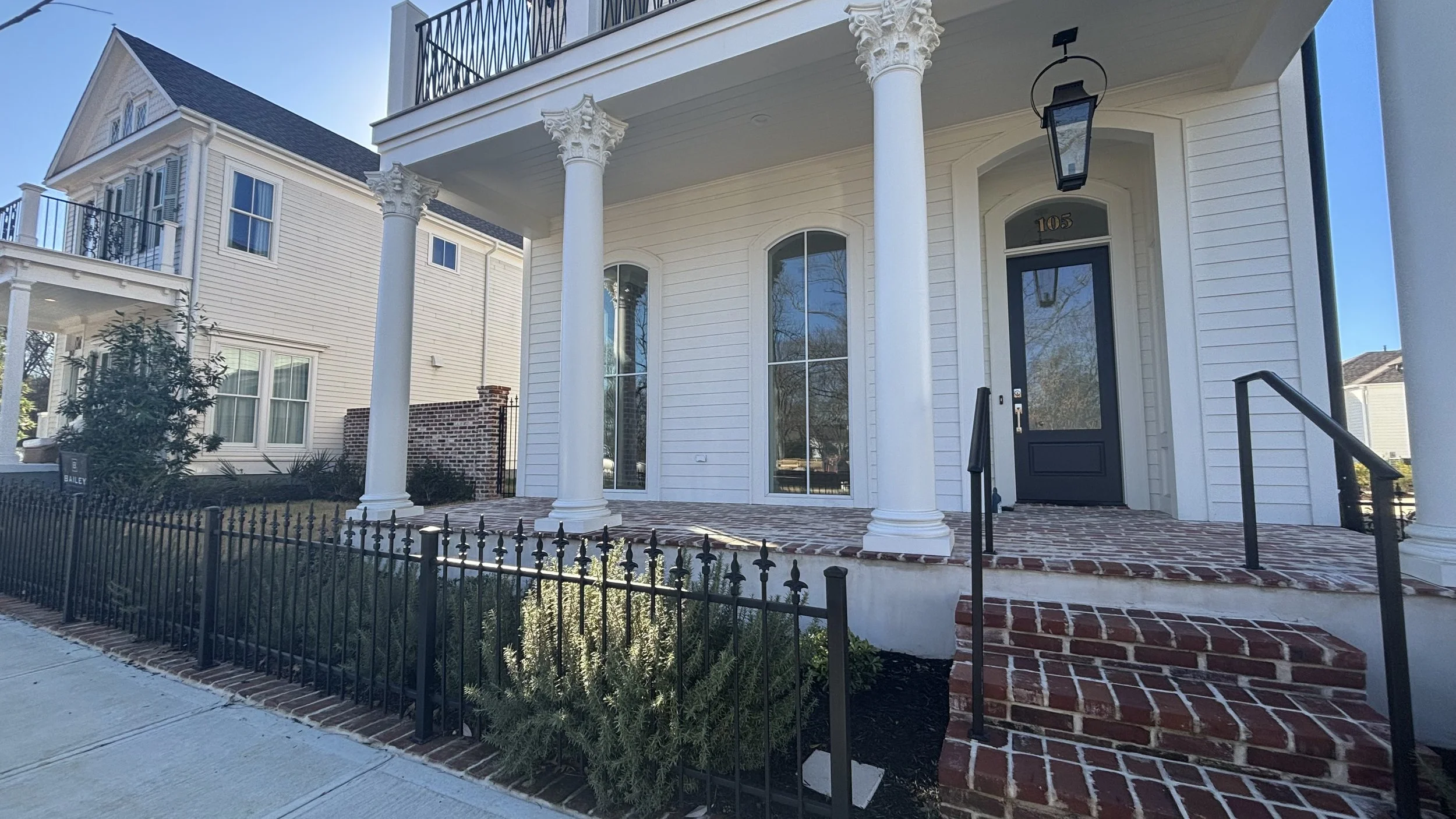 Front view of a white house with a dark door, large arched windows, and four decorative columns on the porch. There is a brick walkway and steps leading to the door, edged by a black metal fence and small landscaped area.