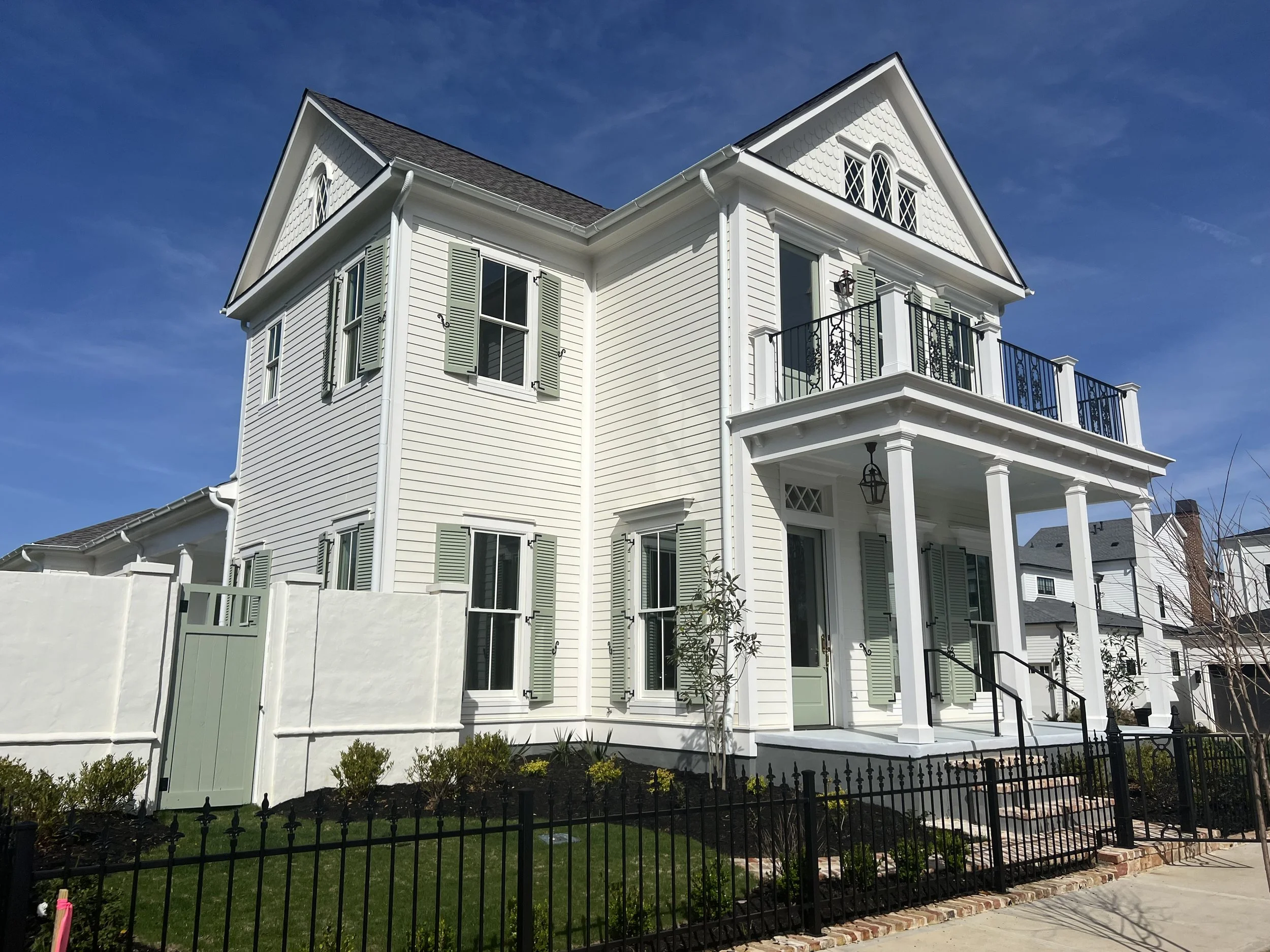 A large, white, three-story house with a front porch, decorative shutters on the windows, and a second-story balcony with black wrought iron railings, under a clear blue sky.