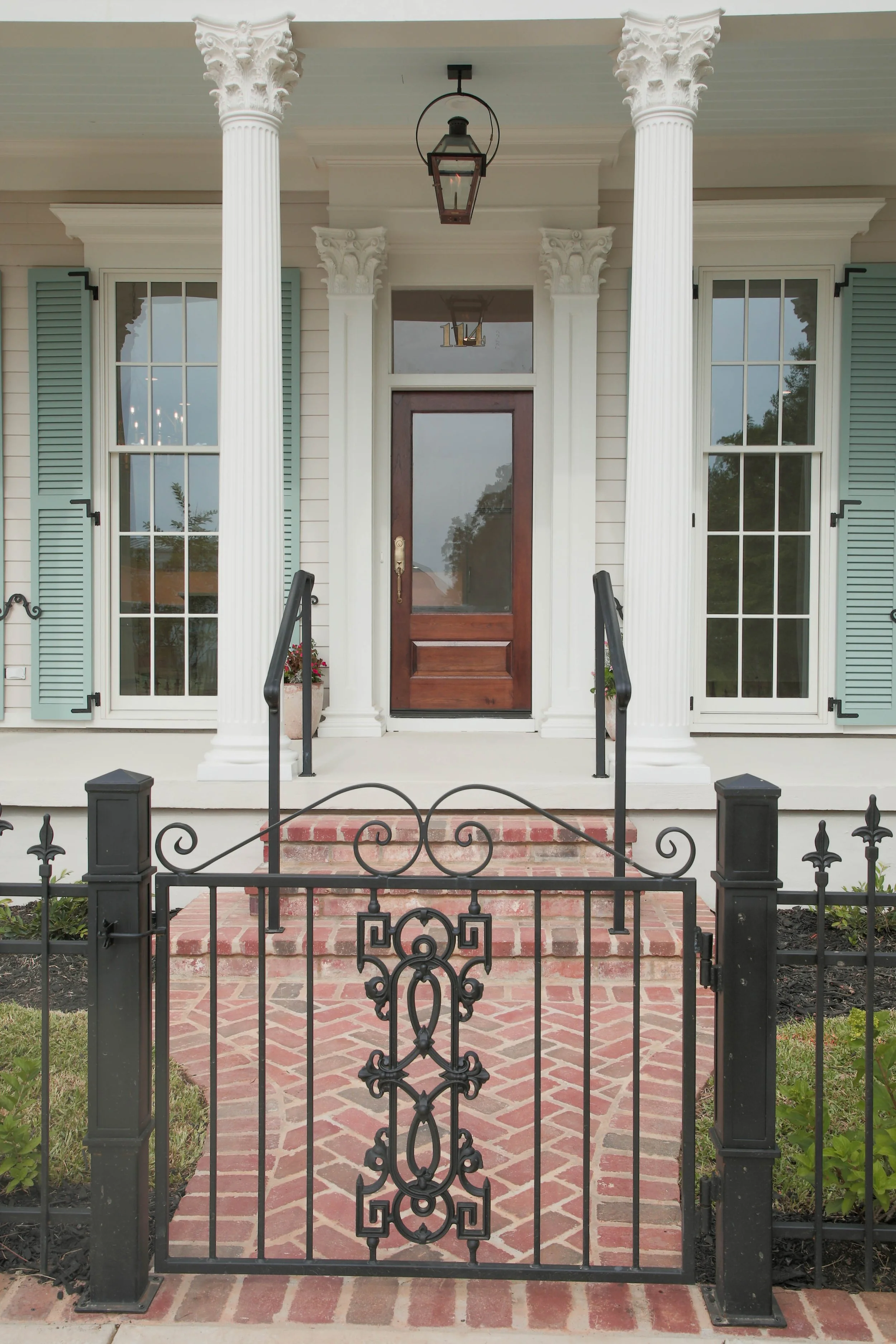 Front porch of a house with brick steps, black wrought-iron fence, white columns, a wooden door, large windows with shutters, and hanging outdoor lanterns.