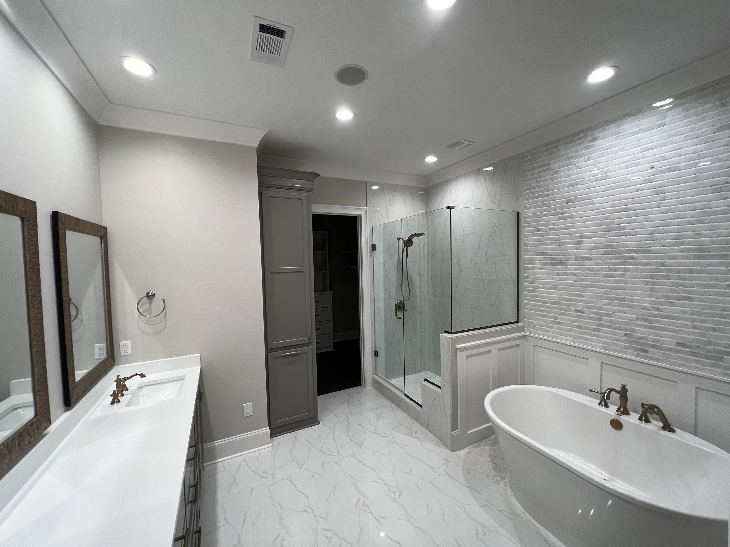 Modern bathroom with white marble floor, white walls, a bathtub with gold fixtures, a separate shower with glass enclosure, and a gray vanity with two mirrors.