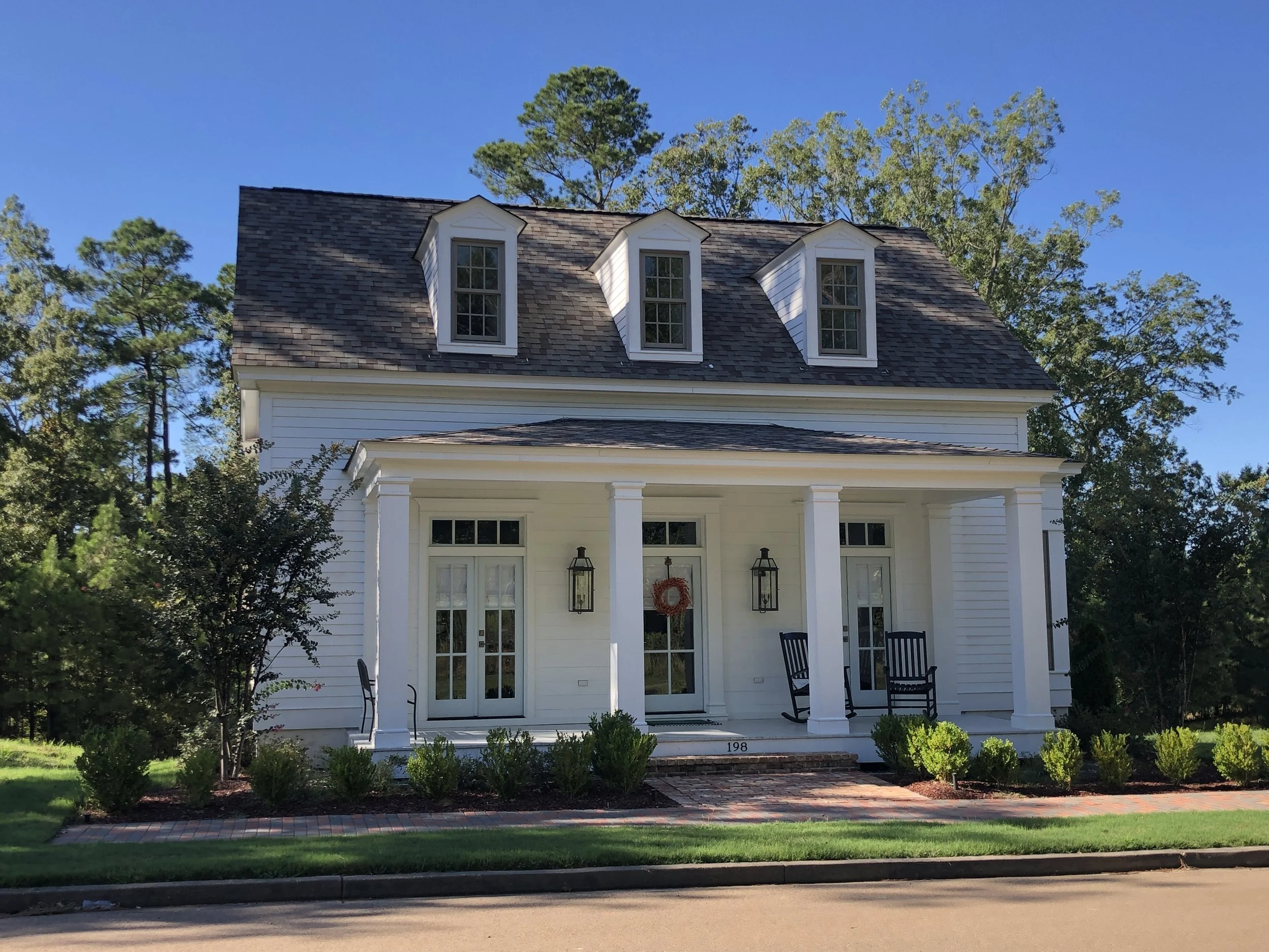 A two-story white house with three dormer windows on the roof, a front porch with four columns, and two black rocking chairs, surrounded by green shrubs and trees under a blue sky.
