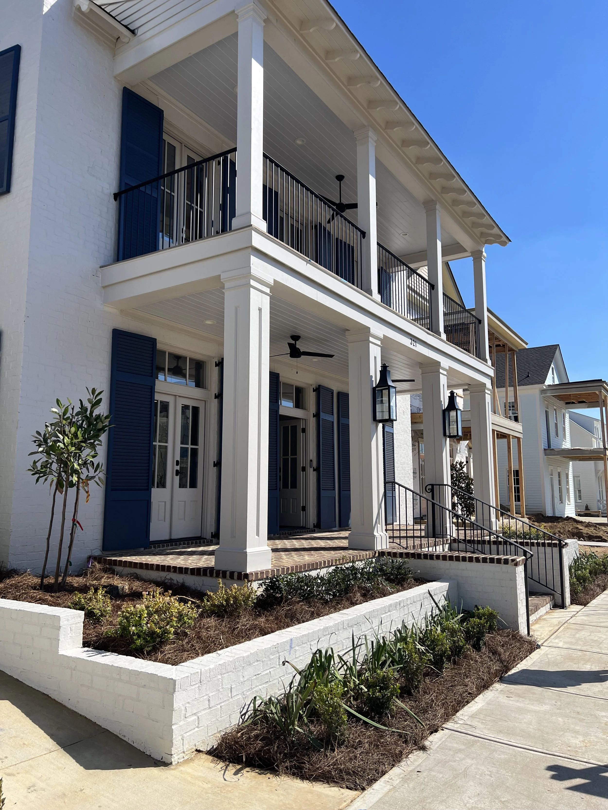 A two-story white house with blue shutters and a front porch with two doors, brick steps, black iron railings, and black outdoor lanterns. There is landscaping with small plants and a tree in front.