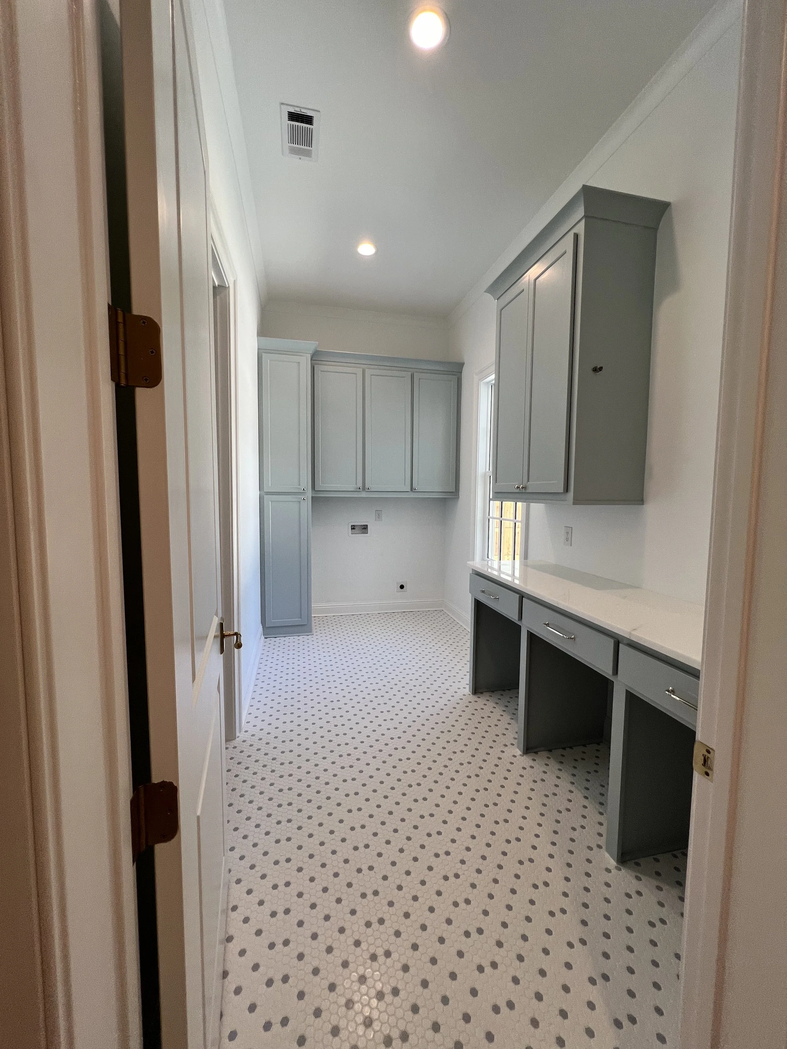 A kitchen with white walls and cabinets, gray upper cabinets, a countertop, small window, and patterned white and black tile flooring.