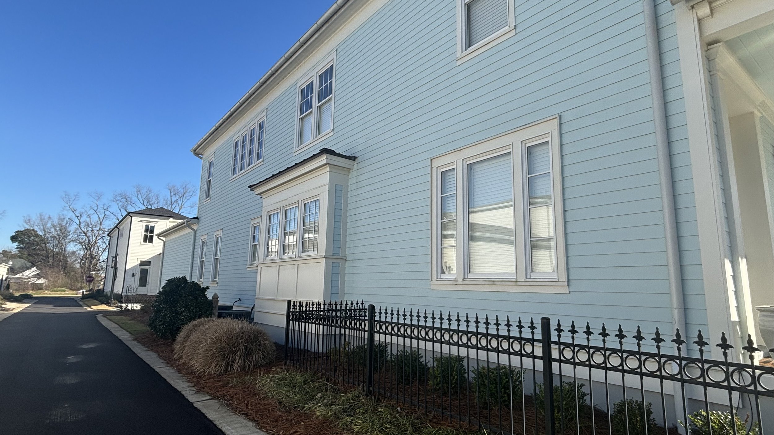 View of light blue residential buildings with white window frames, black wrought iron fence, and landscaped yard on a sunny day.