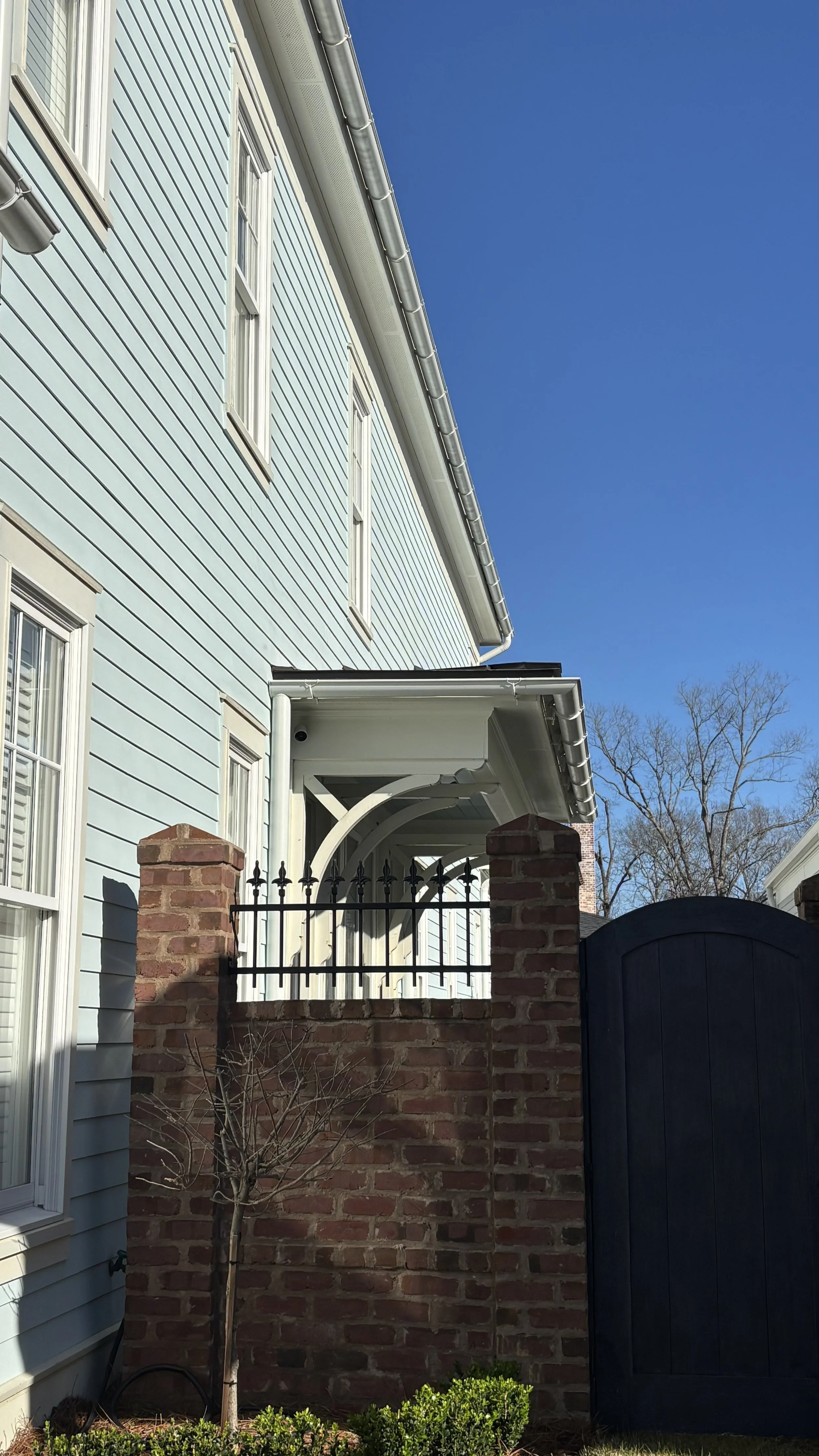 A light blue house with white-framed windows, brick and black wooden fence, and a small leafless tree in front, under a clear blue sky.