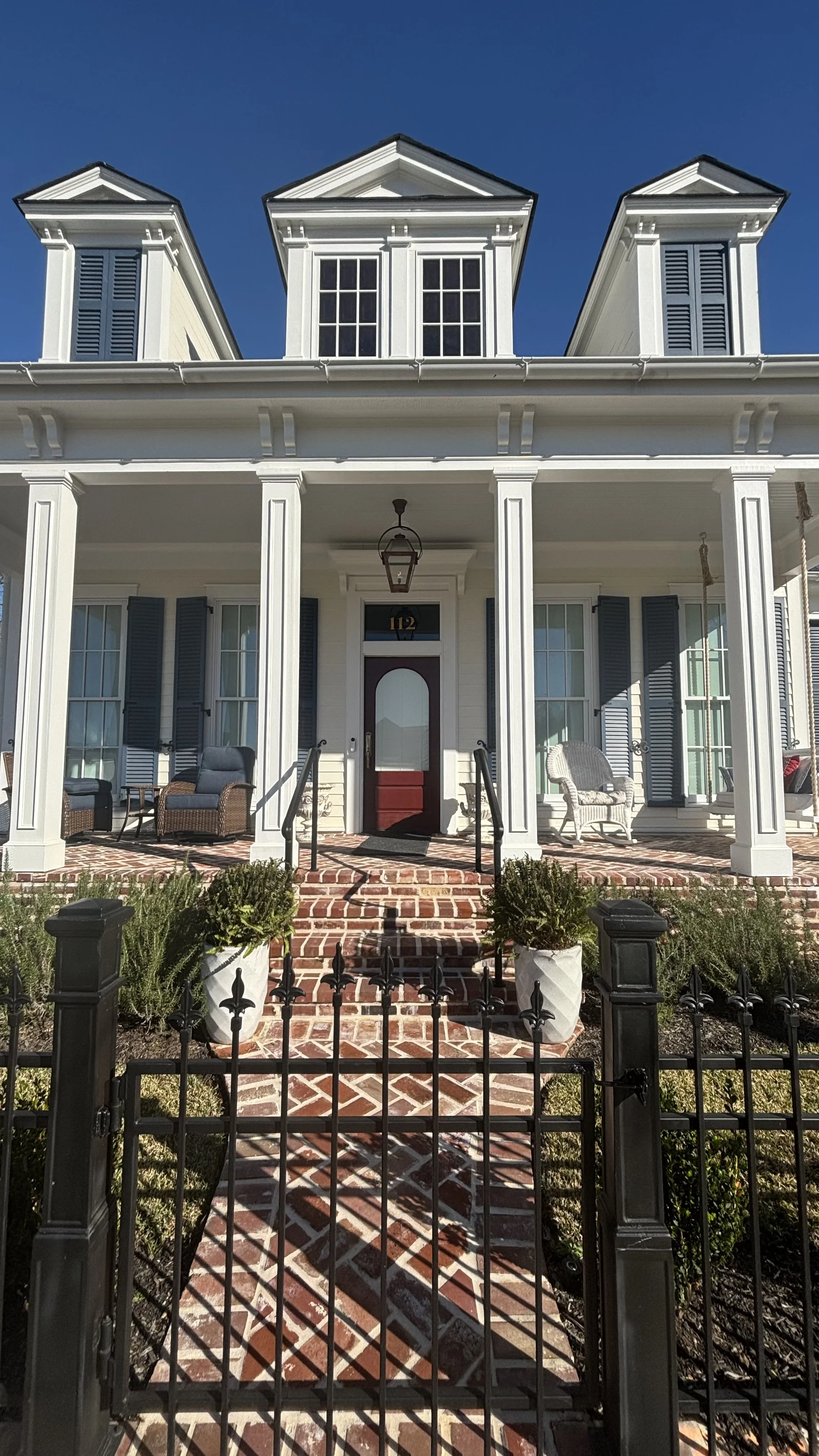Front view of a white, two-story house with a porch, black railings, and a brick walkway leading to the front door. The house has three dormer windows on the roof, black shutters on the windows, and outdoor seating on the porch.