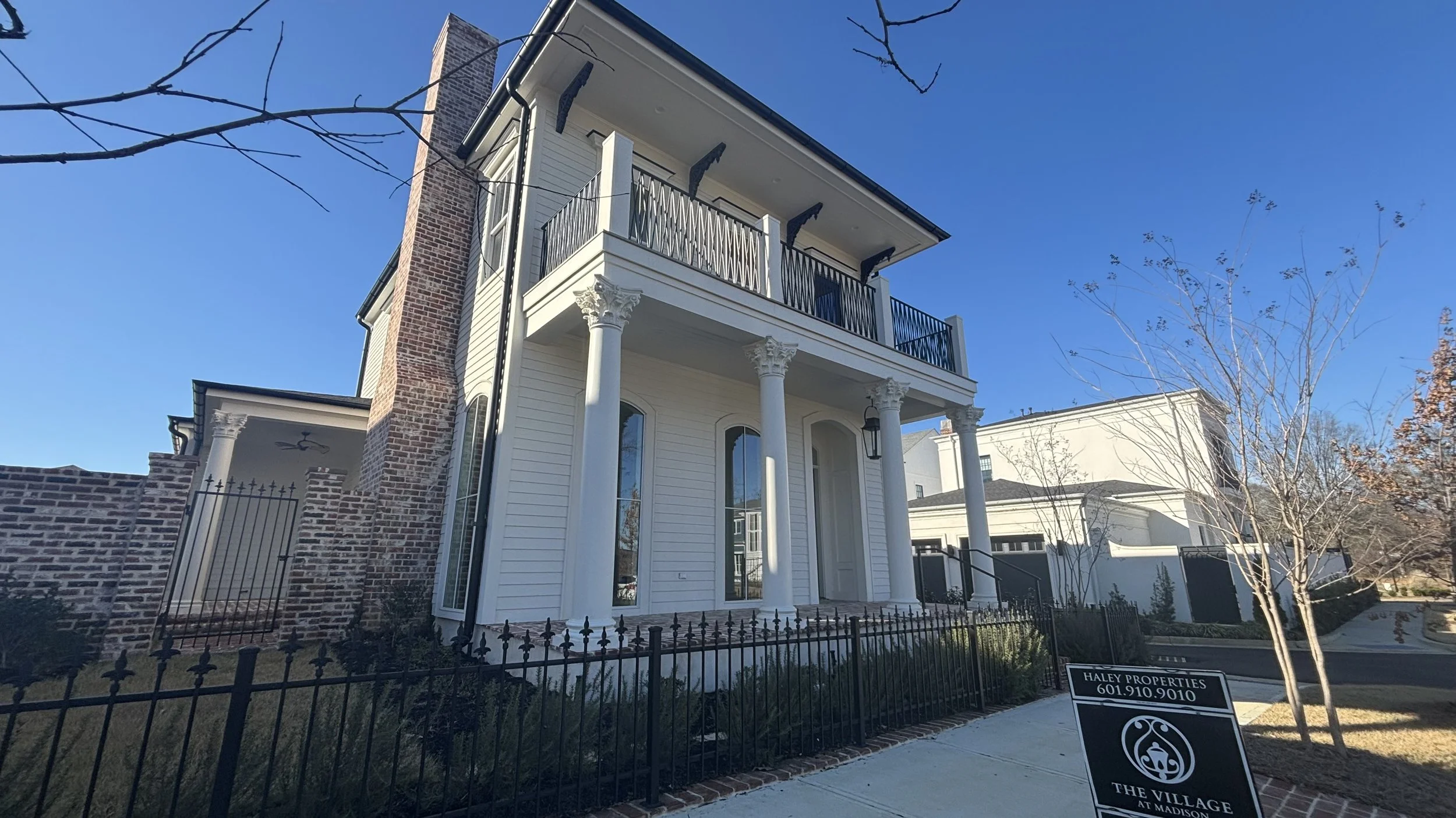 A two-story house with white siding, tall windows, and black balconies, featuring classical columns on the front porch, surrounded by a black metal fence and a garden, with a sign advertising Haley Properties.