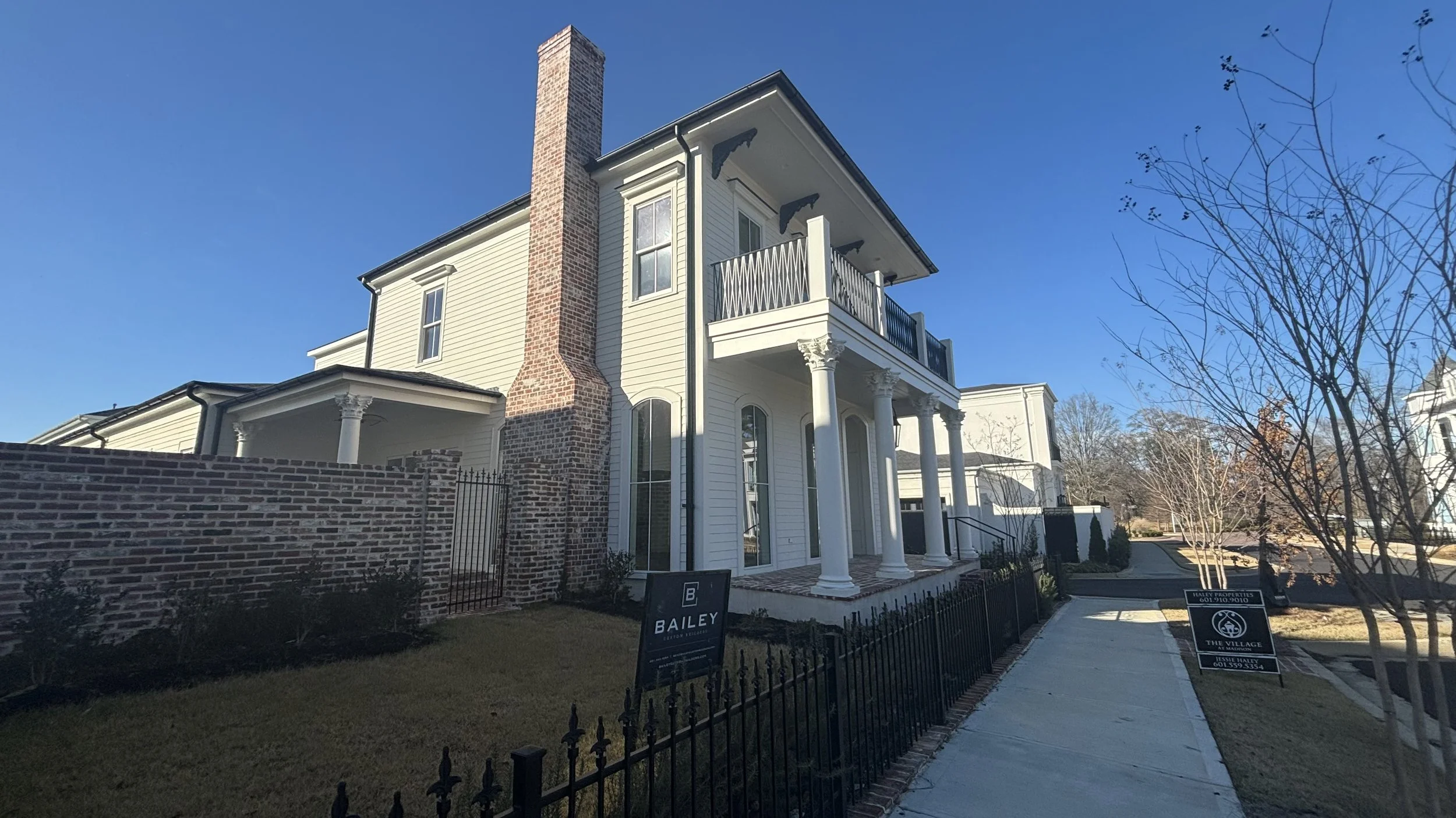 A large, modern multi-story house with a brick chimney, white siding, and decorative columns at the entrance, situated along a sidewalk with signs in the yard. The house has a small balcony on the upper level and is surrounded by a black wrought iron