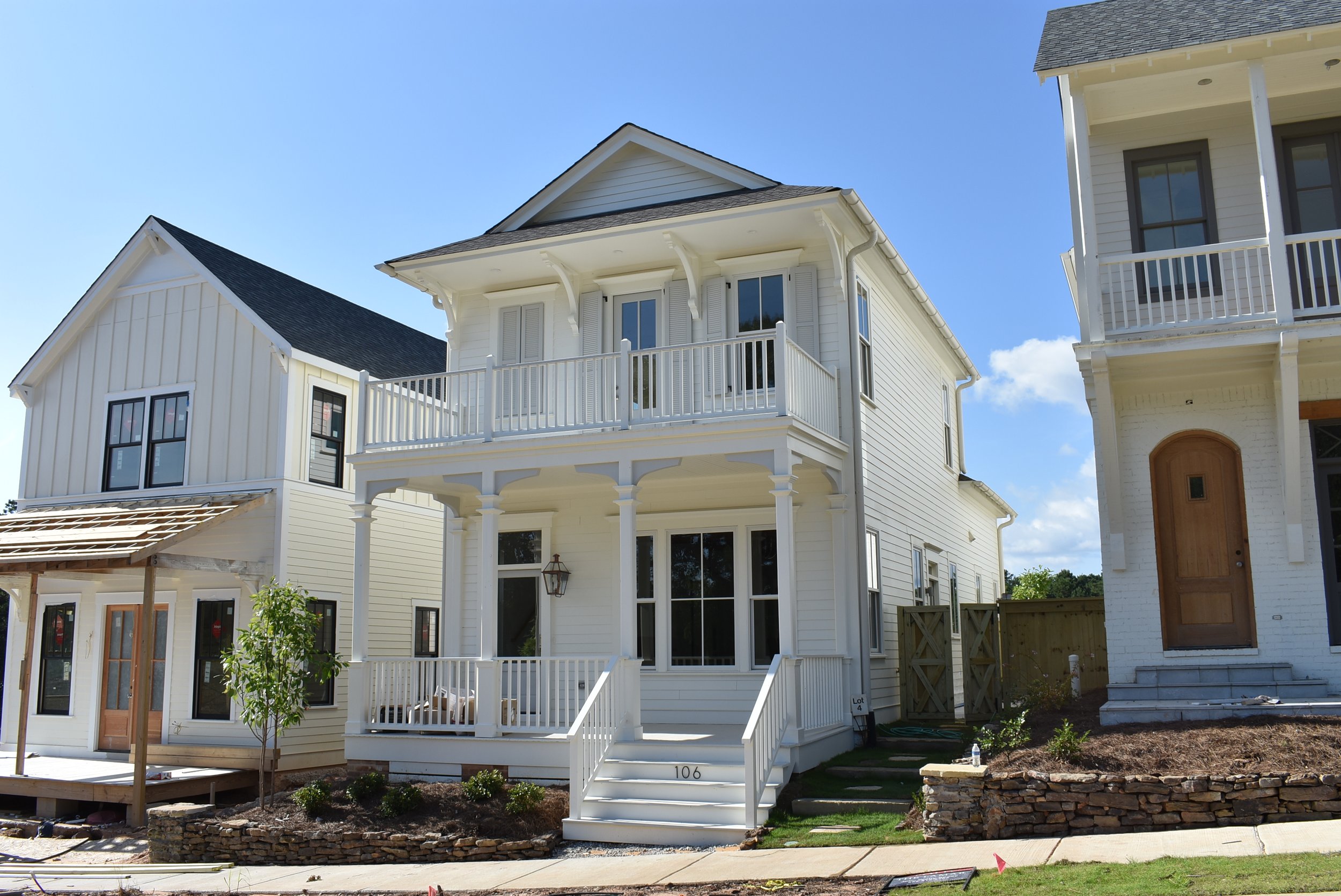 A multi-story white house with a balcony and porch, surrounded by a small yard and neighboring buildings, under a clear blue sky.