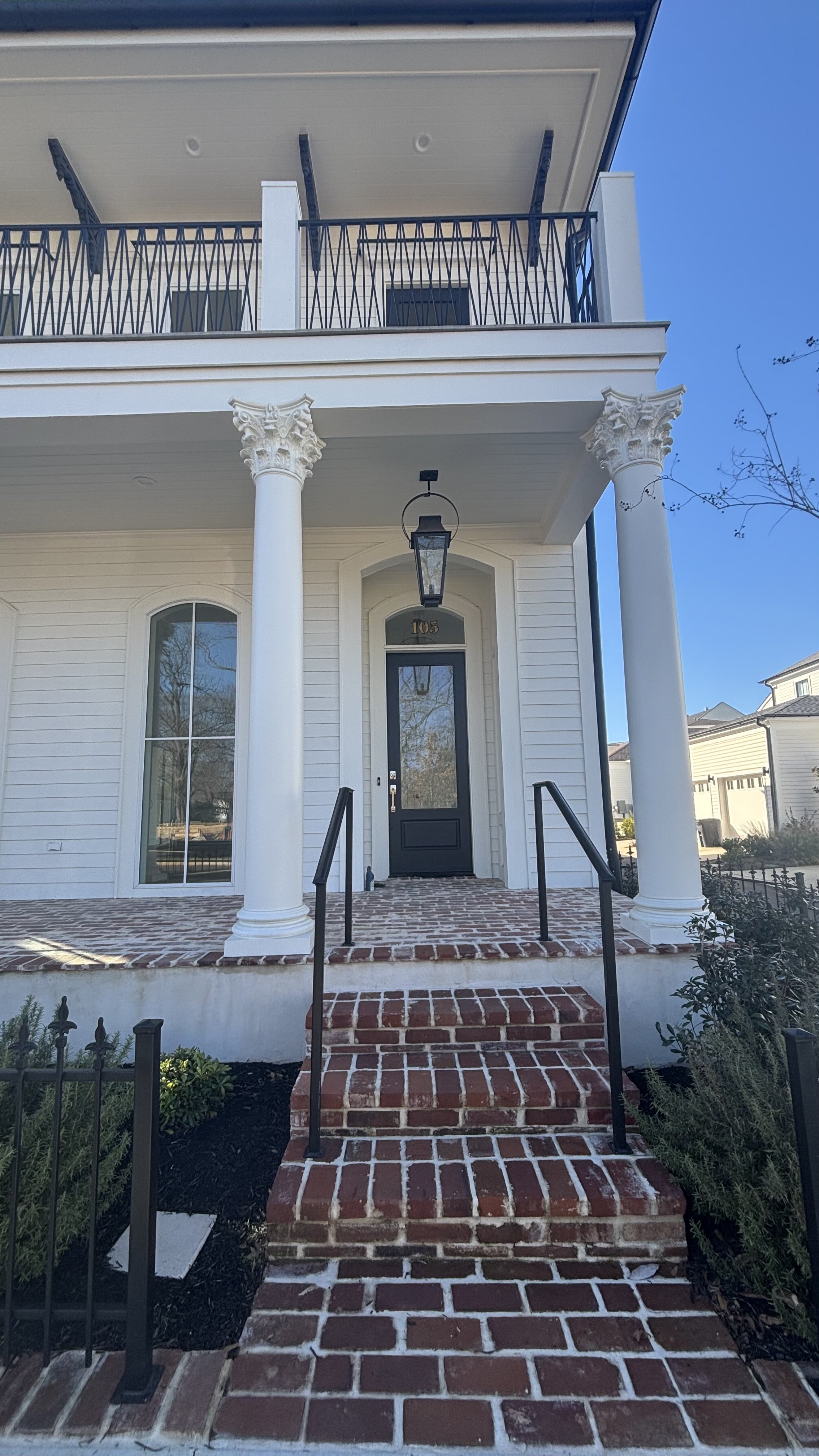 Front view of a two-story house with white siding, brick steps, black railing, and large columns supporting a balcony, with a black door and lantern hanging above