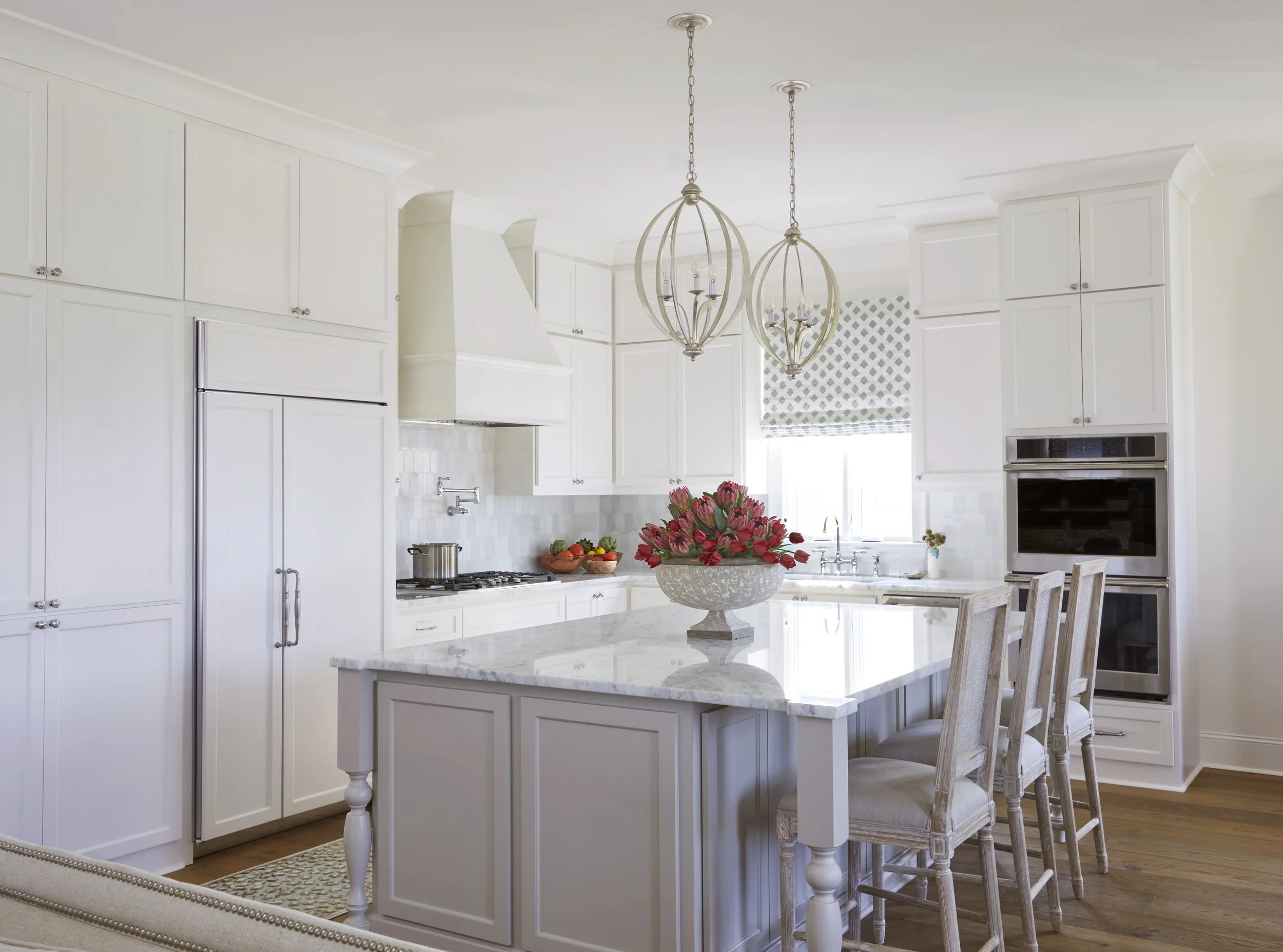 Bright white kitchen with marble island, built-in oven, and stainless steel refrigerator. Light wood chairs around the island and pink flowers in a bowl on top.